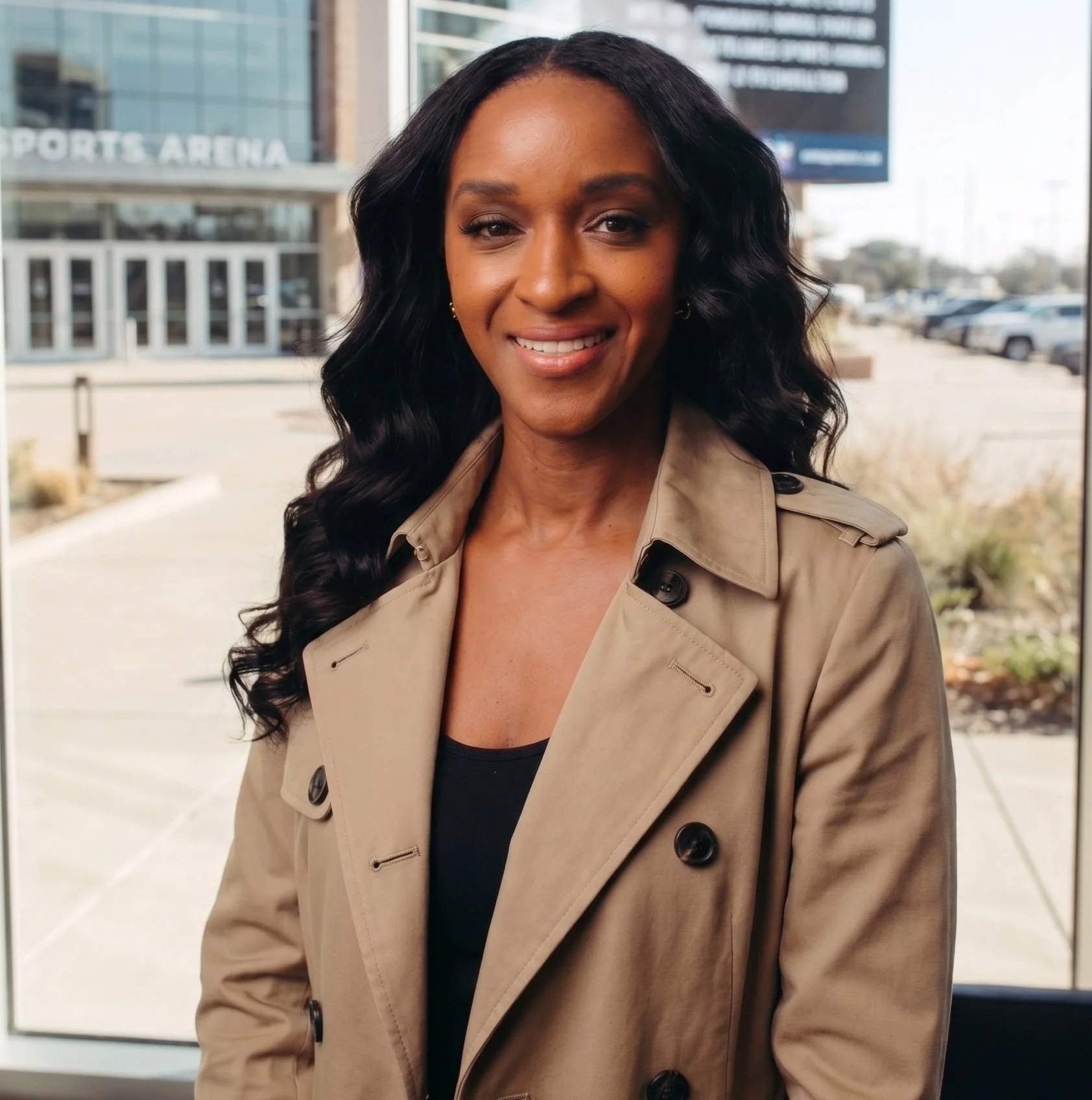A woman with long black curly hair smiling while standing outside in front of a building with glass windows and a parking lot.