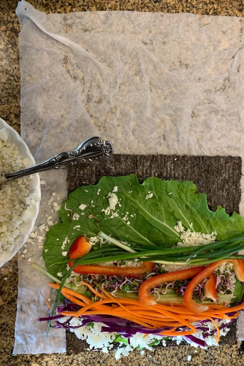 Nori layered with collard greens, cauliflower rice, and colorful vegetables before rolling raw vegan sushi.