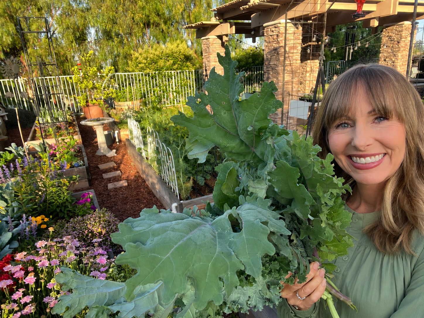 Michelle Stilson, creator of Green Food Crush, smiling in her garden while holding a large bundle of leafy greens beside raised beds and colorful flowers.