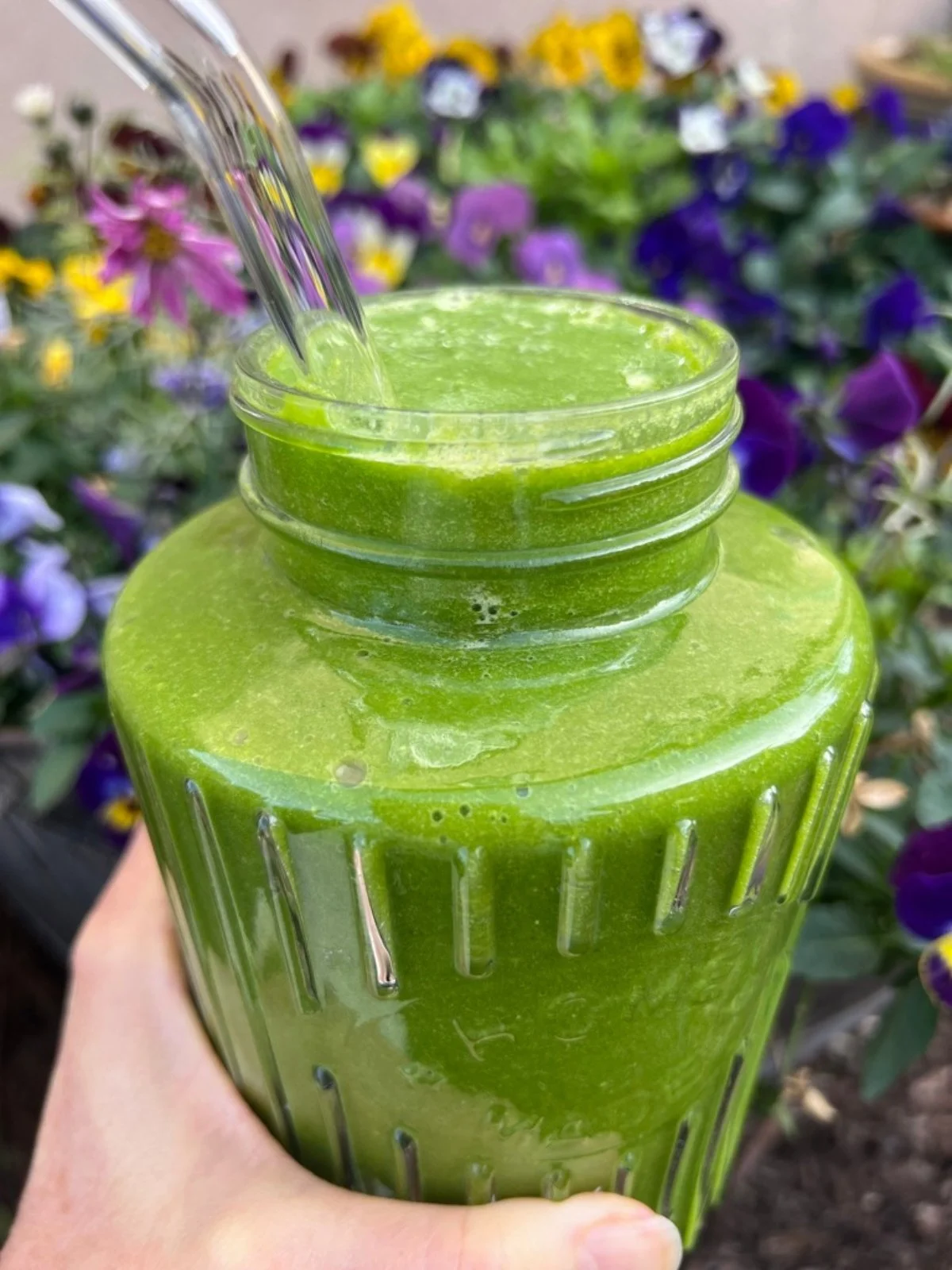 Bright green hyper-nourishing smoothie in a glass jar surrounded by flowers in the garden.
