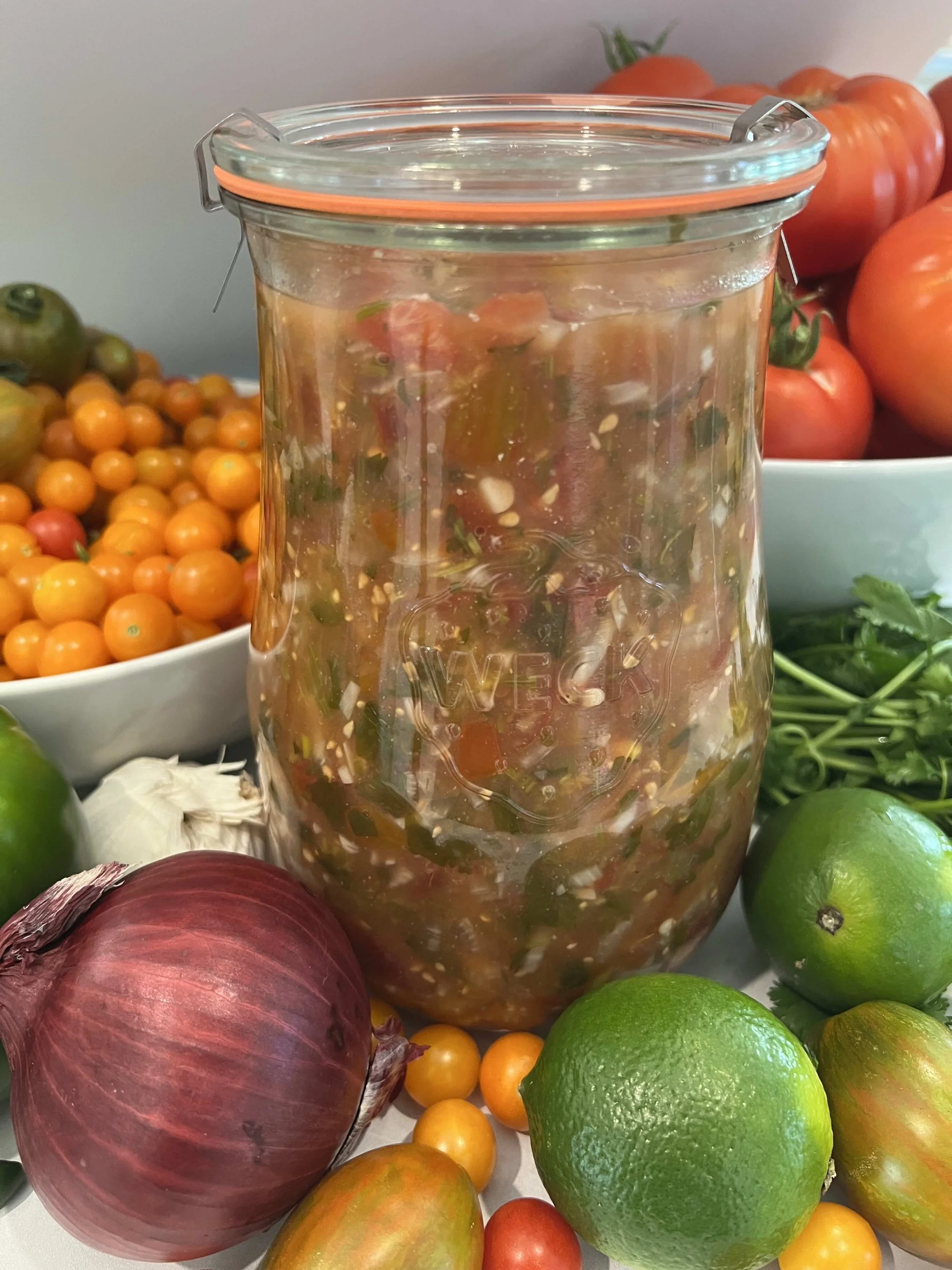 Homemade fermented salsa in a glass jar surrounded by fresh garden tomatoes, lime, red onion, and cilantro.