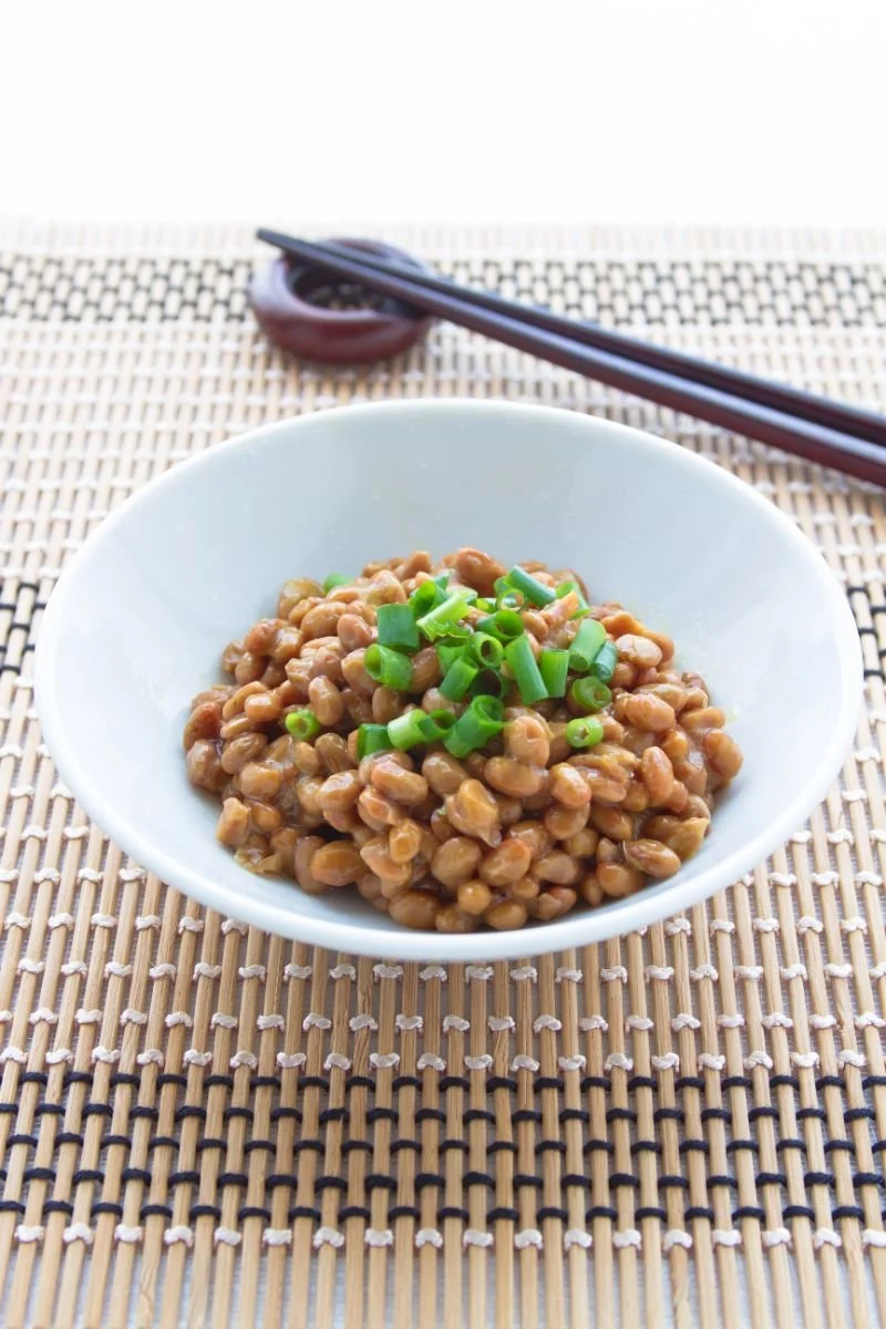 Bowl of natto with spring onions on top with a pair of wooden chopsticks resting nearby.