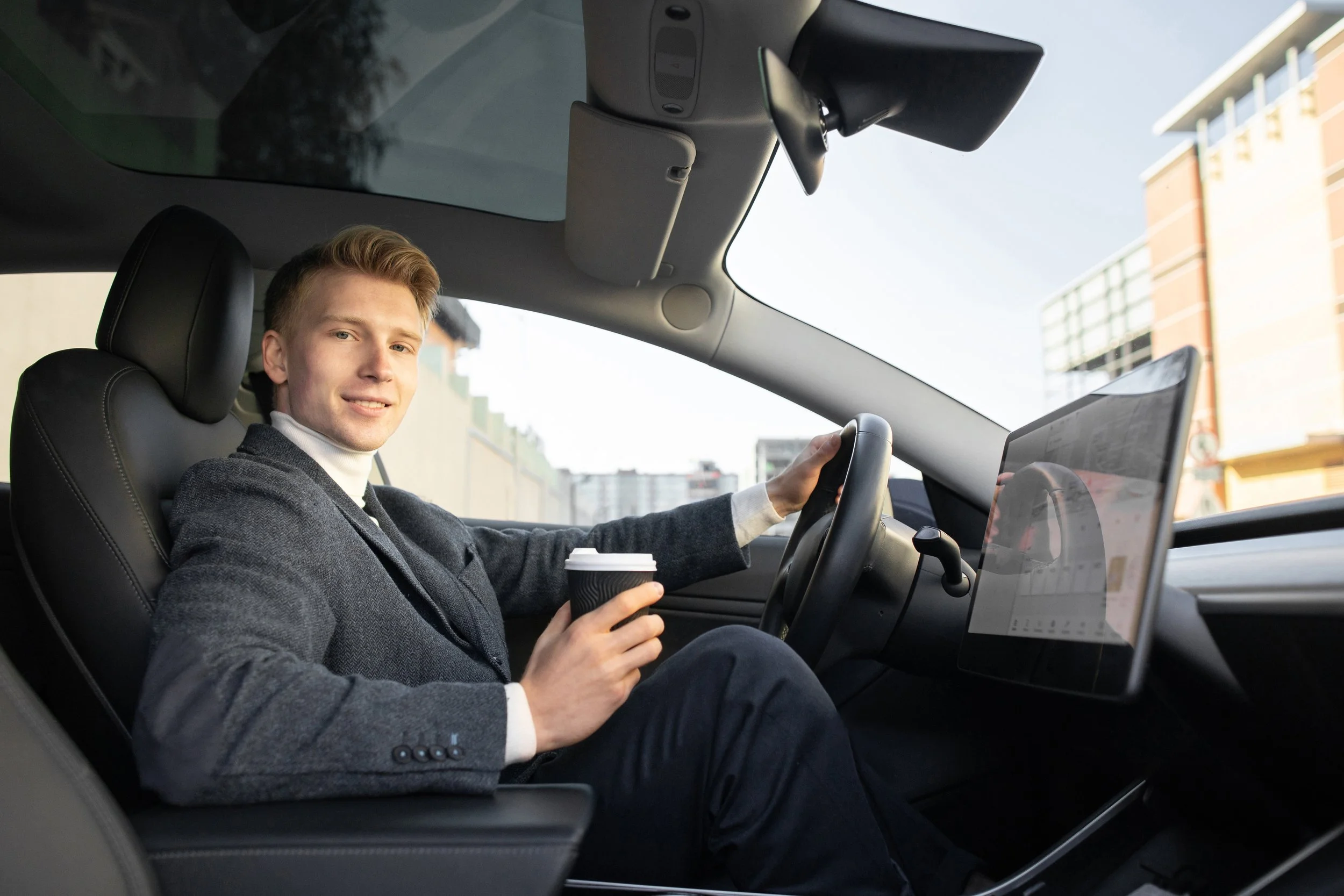 A young man with blond hair, dressed in a gray blazer and white turtleneck, is sitting in the driver's seat of a modern car. He is smiling and holding a coffee cup in his left hand, while his right hand is on the steering wheel. The interior of the car is equipped with a large screen on the dashboard. Outside, there are buildings and a clear sky.