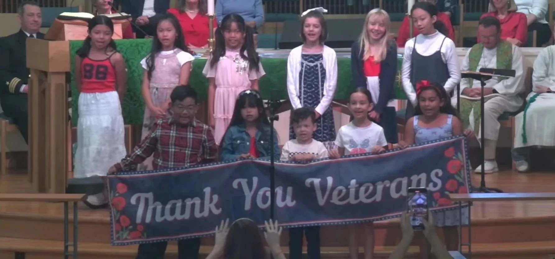 Group of children on stage holding a banner that reads 'Thank You Veterans' during a ceremony or event.