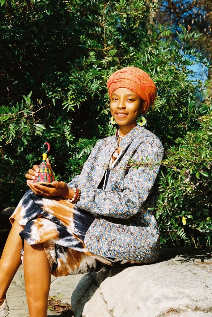 A woman sitting outdoors on a stone, surrounded by green bushes, wearing a patterned blazer, tie-dye skirt, orange head wrap, and colorful earrings, holding a beaded bag and smiling.