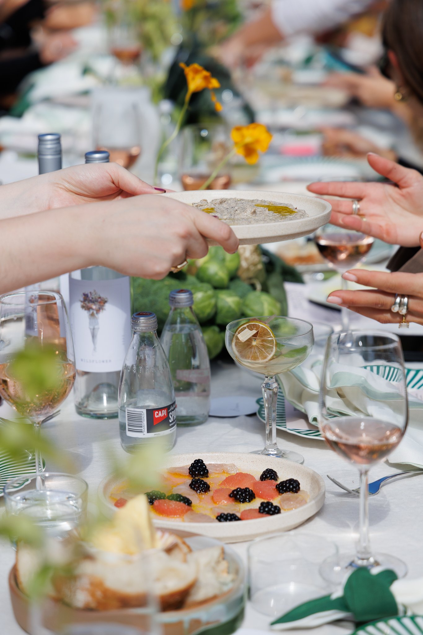 Close-up of a table set for a meal with a hand holding a small dish, surrounded by glasses of wine, bottles, a fruit plate with blackberries, a lemon slice, and a catering scene with people toasting in the background.
