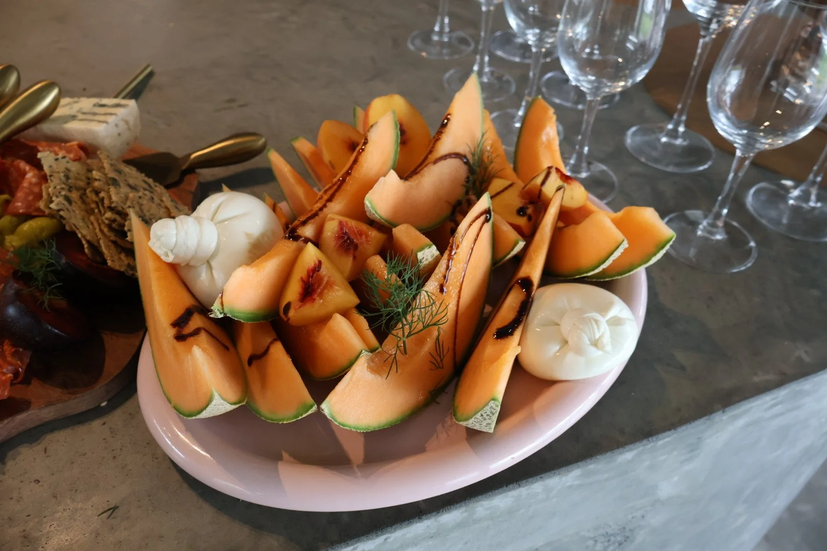 A white oval plate filled with sliced cantaloupe melon topped with chocolate sauce, garnished with sprigs of fresh dill, and accompanied by two large dollops of cream. In the background, there are empty wine glasses on a table.
