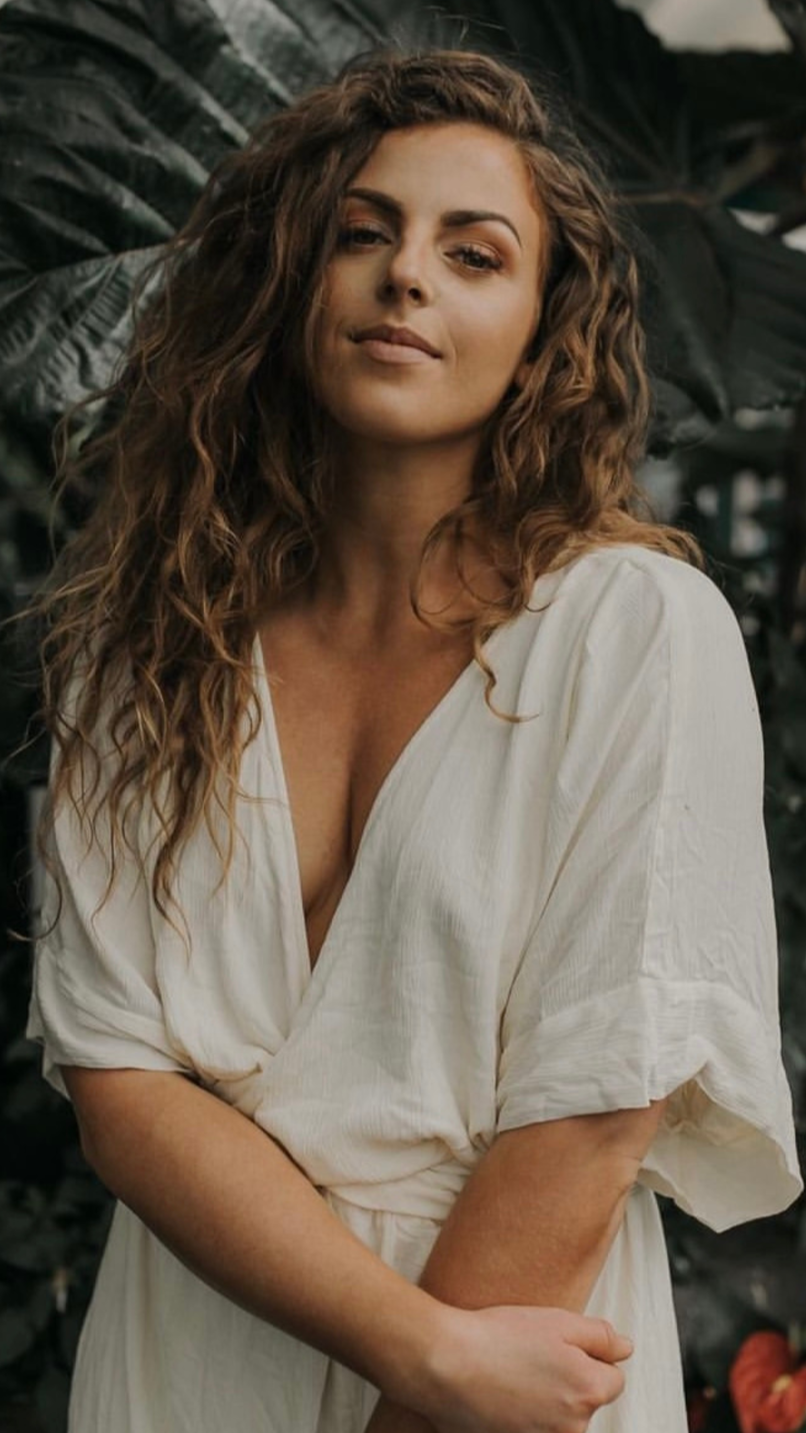 A woman with long, curly brown hair wearing a white dress with a deep V-neck, standing outdoors with dark green foliage in the background