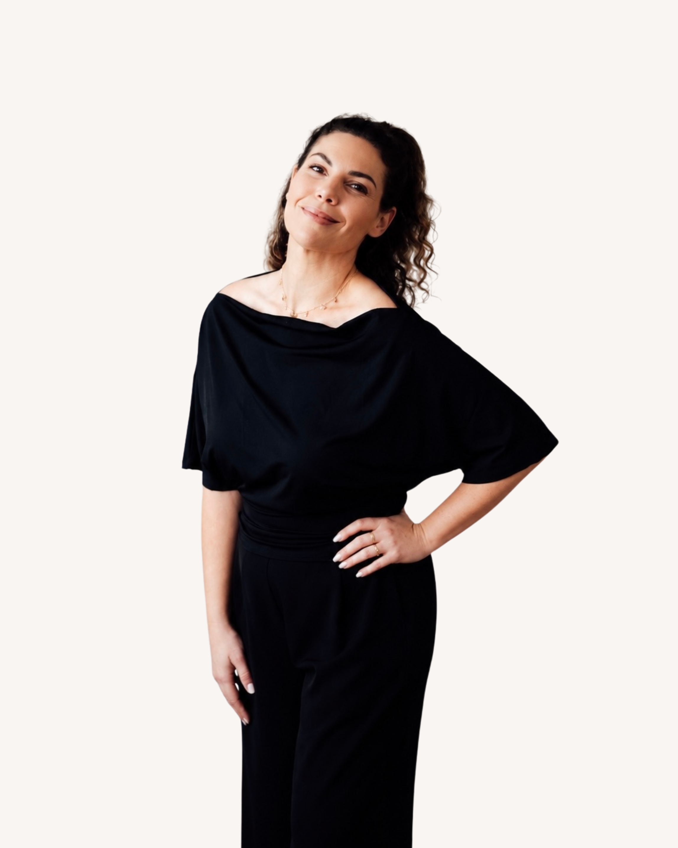 A woman with dark, curly hair wearing a black off-the-shoulder dress and a gold necklace, posing with one hand on her hip against a plain backdrop.
