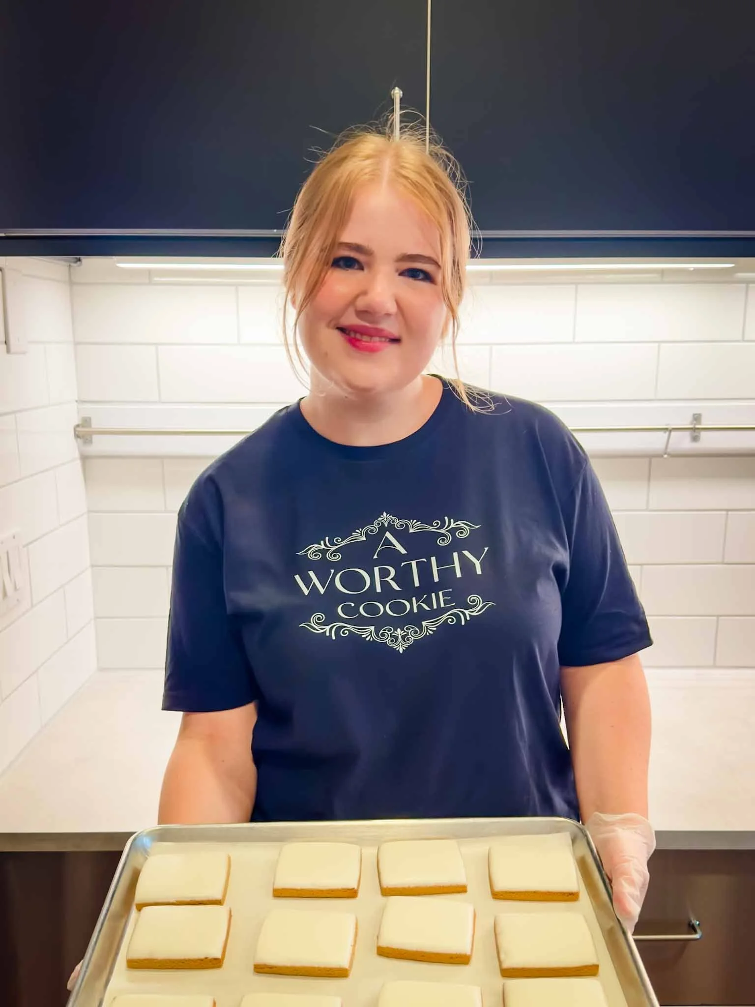 A young woman with red hair smiling and holding a tray of square cookies in a kitchen.