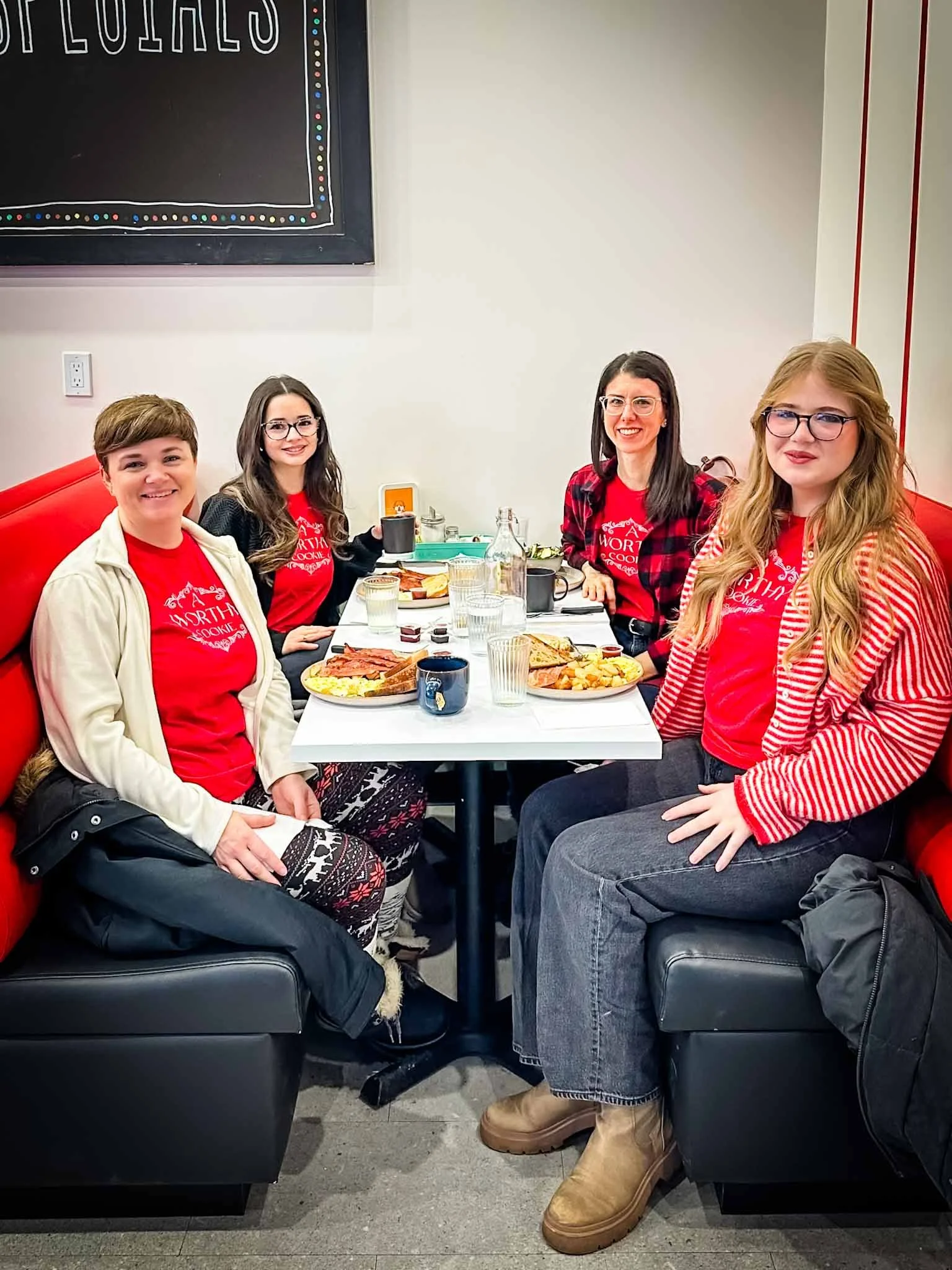 Four women sitting at a restaurant table with breakfast food, smiling at the camera, wearing red shirts with white writing, with a chalkboard on the wall behind them.