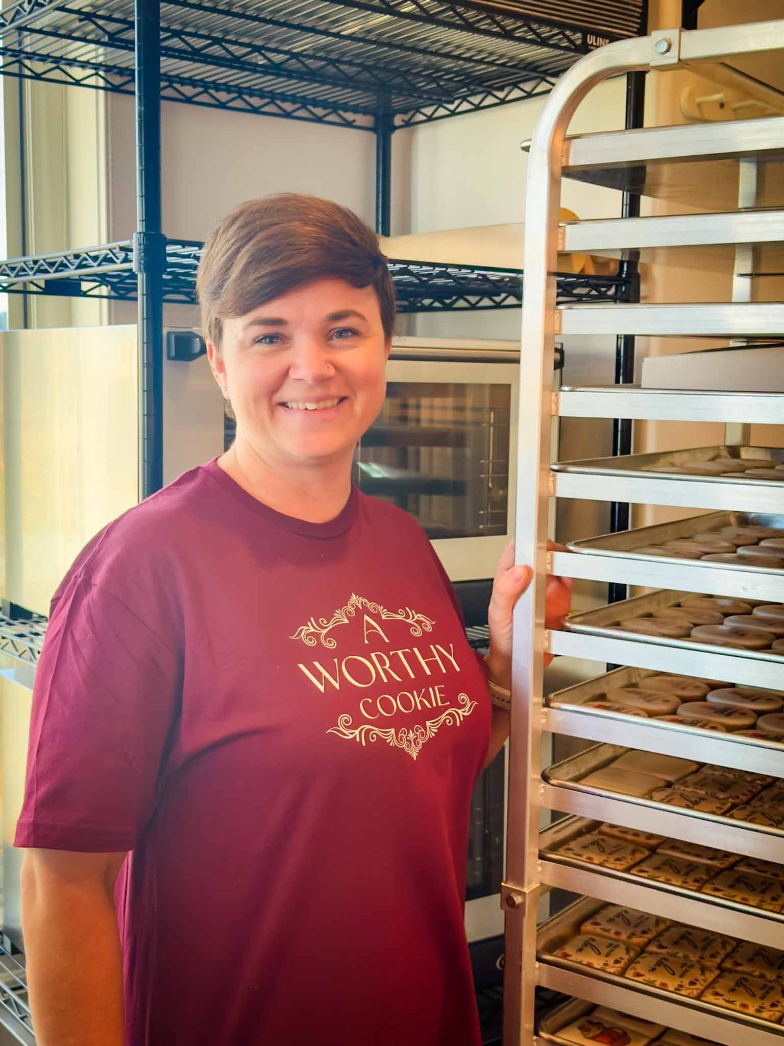 Woman with short brown hair smiling, wearing a maroon T-shirt that says "A WORTHY COOKIE" in decorative font, standing next to a metal rack with trays of decorated cookies.