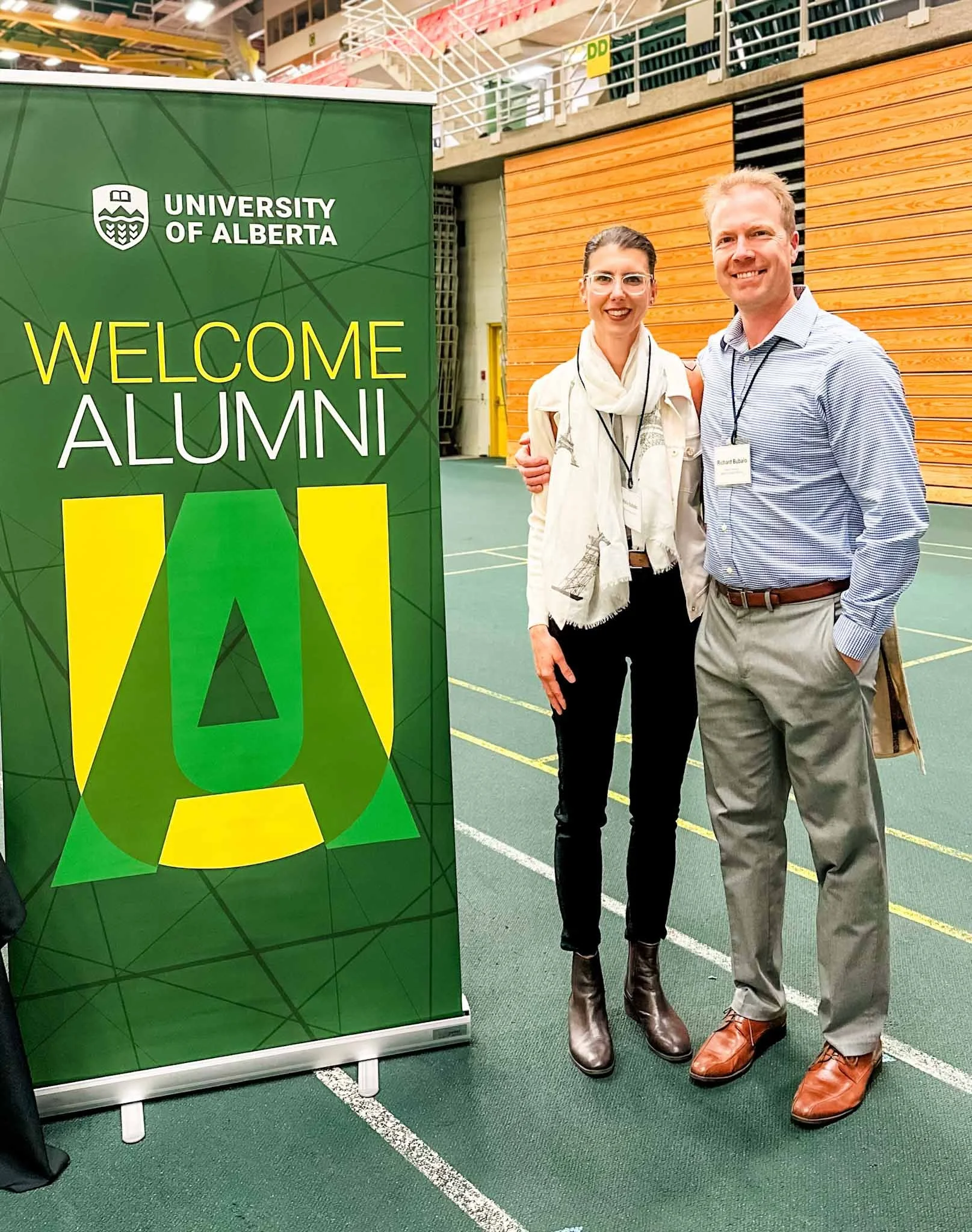 Two people standing indoors next to a green sign that says "Welcome Alumni" from the University of Alberta. The woman on the left is wearing glasses, a white scarf, a beige jacket, black pants, and brown boots. The man on the right has a name tag and