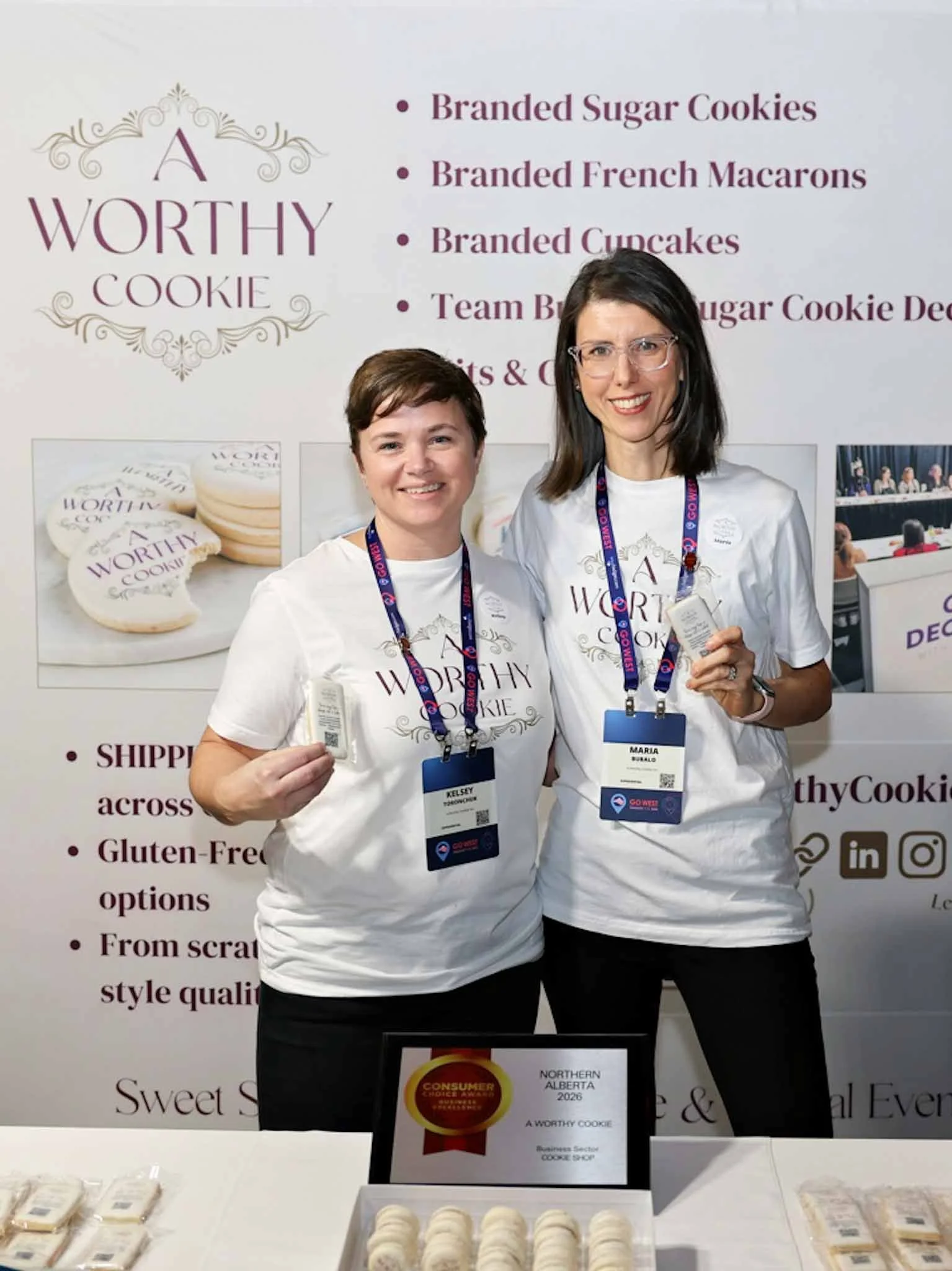 Two women at a tradeshow event, smiling and holding cookies in front of a large sign listing various cookies and desserts, wearing event badges and white T-shirts that say "A WORTHY COOKIE."