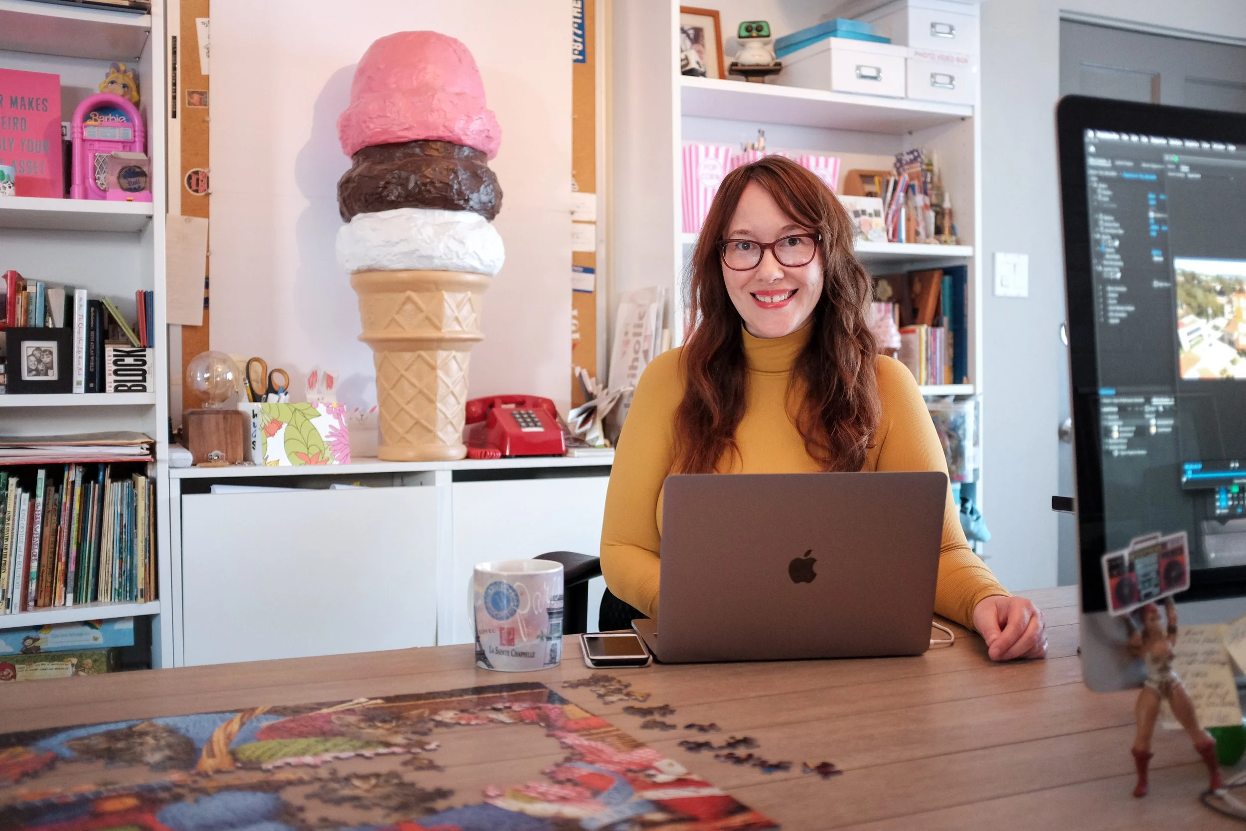 A woman sits with her laptop open in a room that's decorated in a fun way. There is a giant ice cream cone in the background and a puzzle on the table..