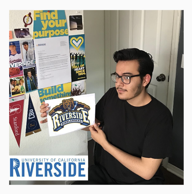  A person holds up a UC Riverside sticker. Behind them is a wall decorated with pennants and their acceptance letter. 
