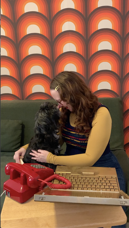 A little dog gives a lady a tiny kiss on her nose. There is fun wallpaper in the background and an old-fashioned phone and laptop in the foreground.