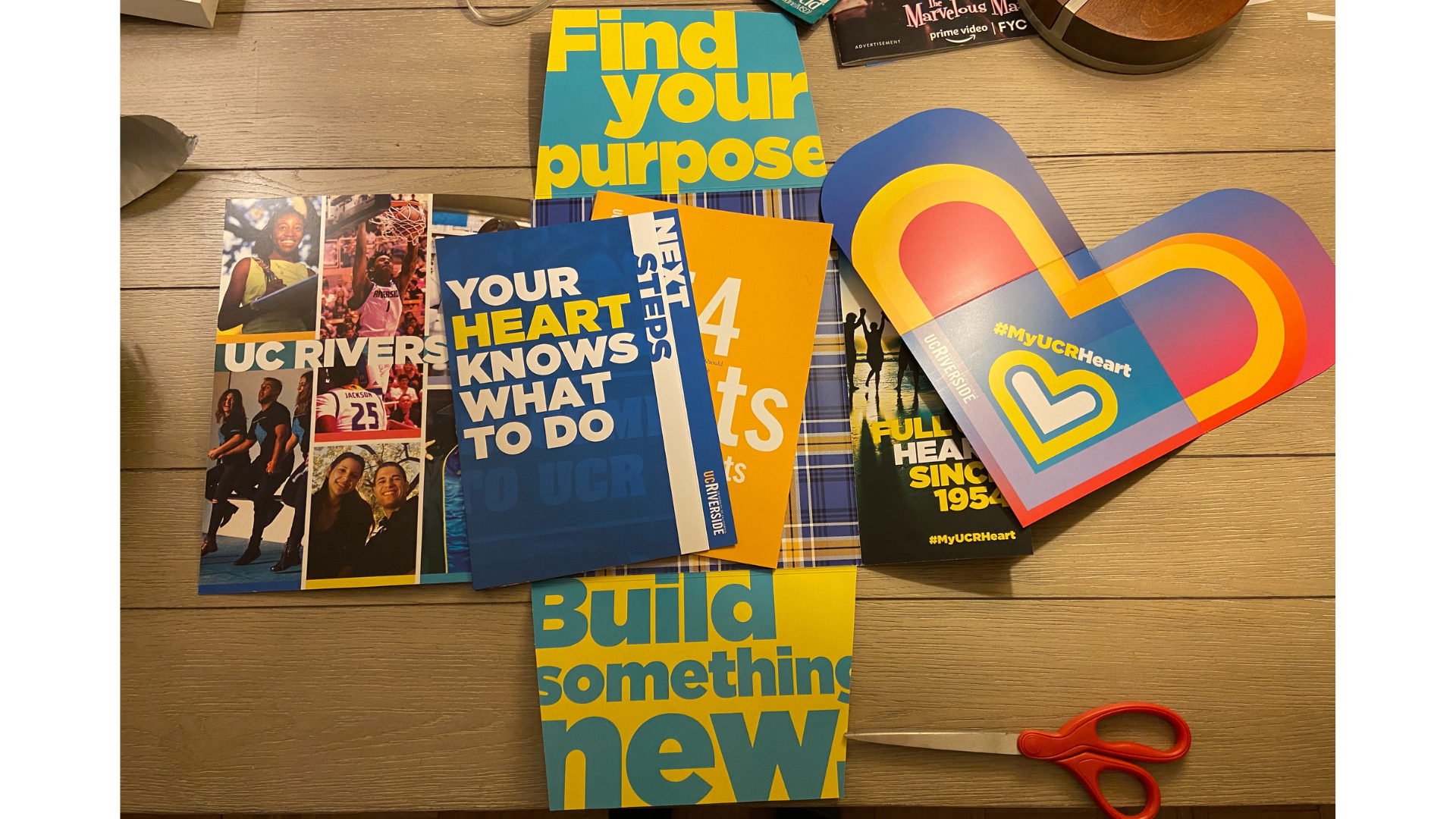 A UCR acceptance packet is open and its contents are spilling out. There are photos of students having fun. Panels say find your purpose and build something new. A pamphlet says your heart knows what to do. There is a big paper heart with #myucrheart
