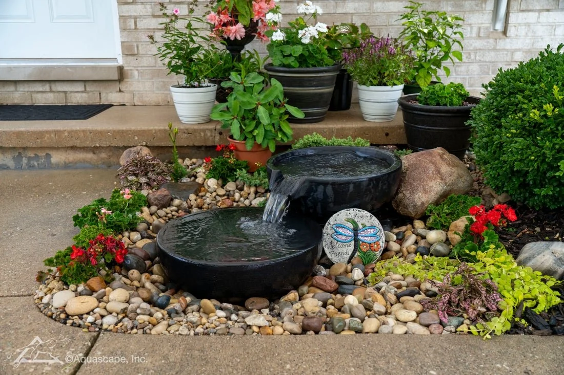 A small garden with two black stone water fountains surrounded by colorful flowers and green plants, with potted plants placed on a concrete porch in front of a house with a brick wall and a white door.