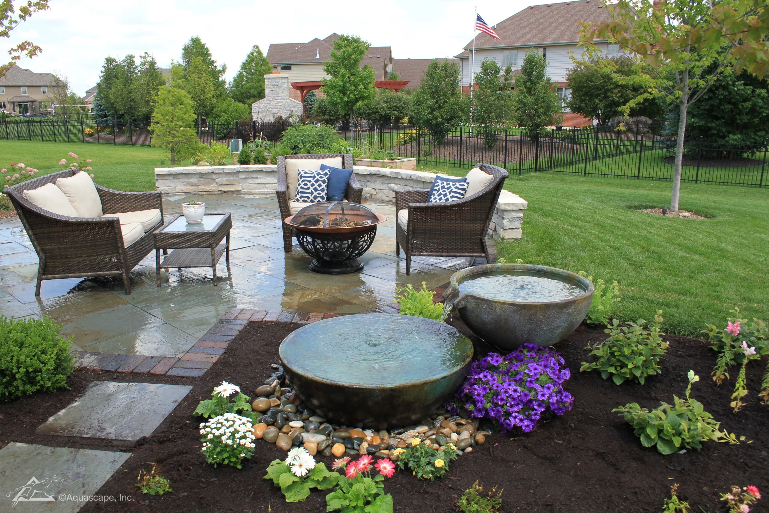 A backyard patio with four outdoor chairs and a table, water features made of large bowls, surrounded by flower beds, trees, and a lawn with a house, a stone chimney, and an American flag in the background.