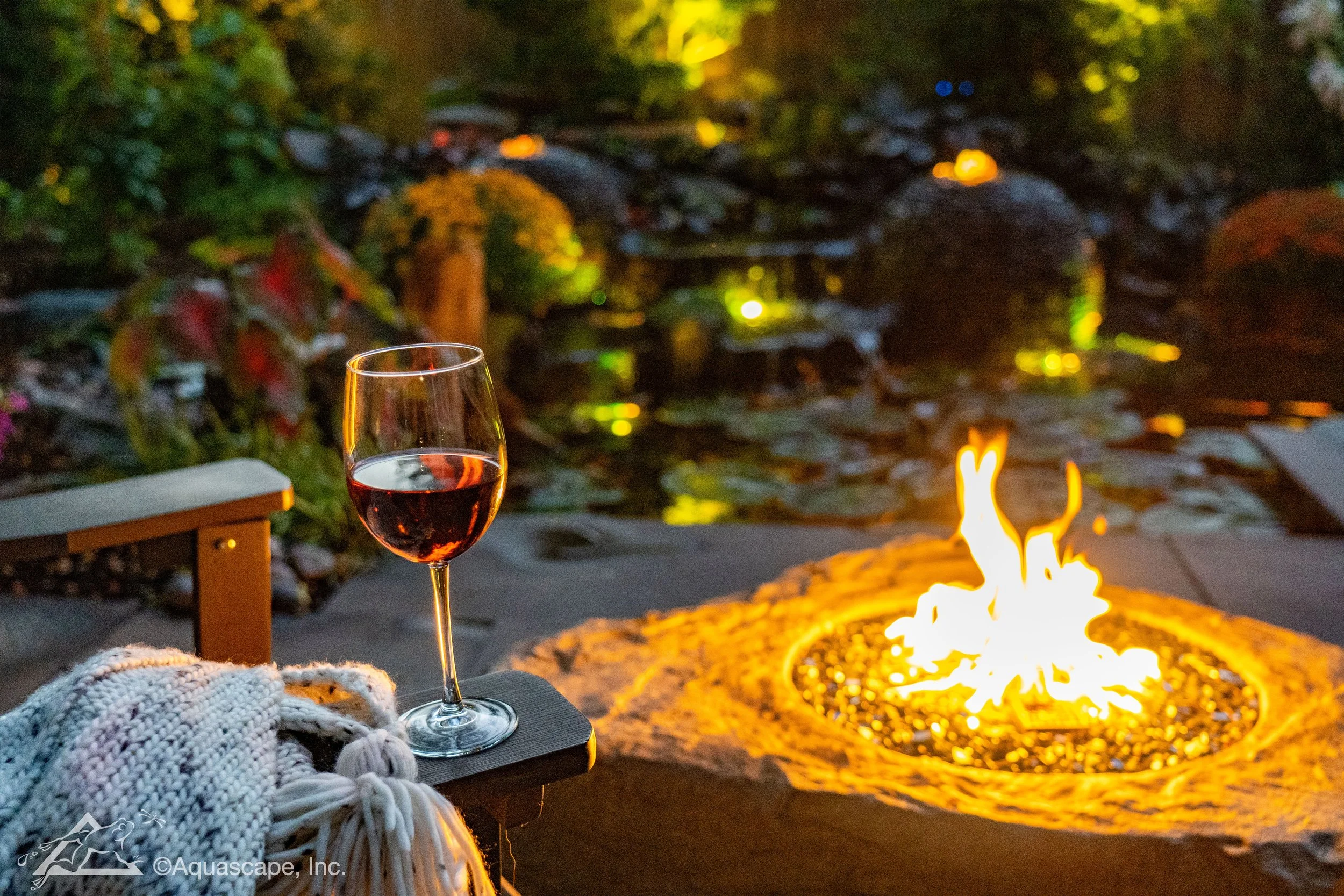 A glass of red wine on a small table with a cozy blanket, outdoors at dusk by a fire pit, with a garden and water feature in the background.