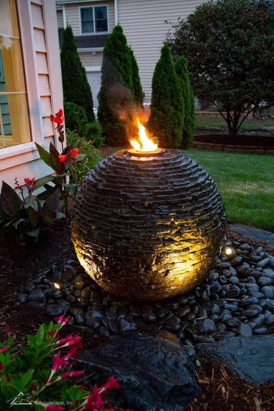 Decorative garden fountain with a flame on top, surrounded by black rocks and plants, in a backyard setting at dusk.