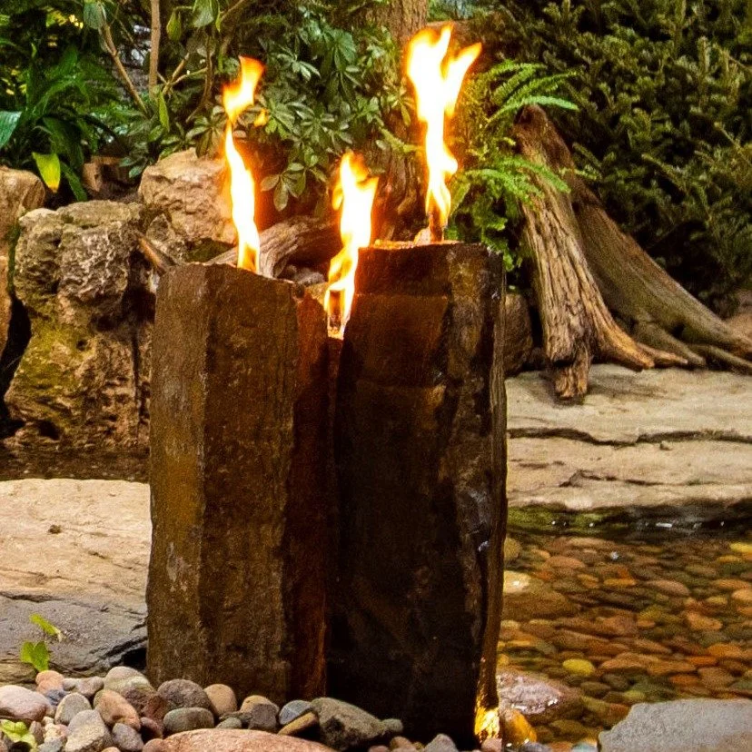 Three pillars. Two elements. One unforgettable focal point. 🔥💧

This custom water feature features three natural basalt columns grouped together, water cascading from the tops while fire bowls glow above &mdash; creating a dramatic contrast that co