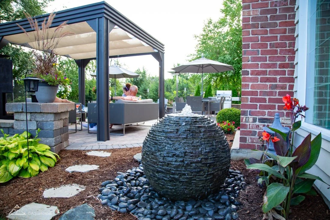 Backyard patio with seating area, umbrellas, and a water fountain surrounded by plants and flowers.