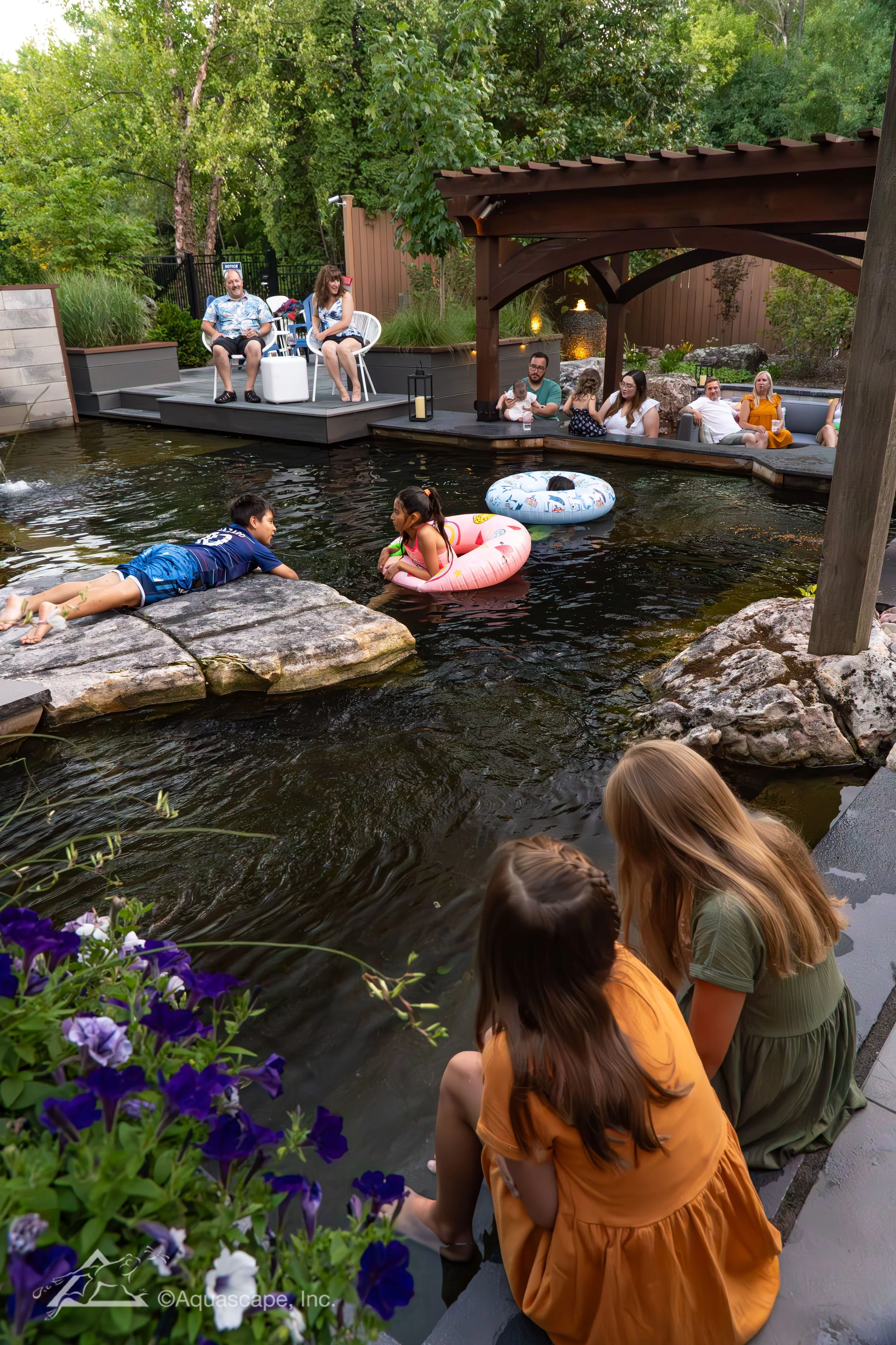 Children and adults enjoying a backyard pond with sitting and swimming areas, including children in swim rings and a boy lying on rocks, surrounded by trees, plants, and outdoor furniture.