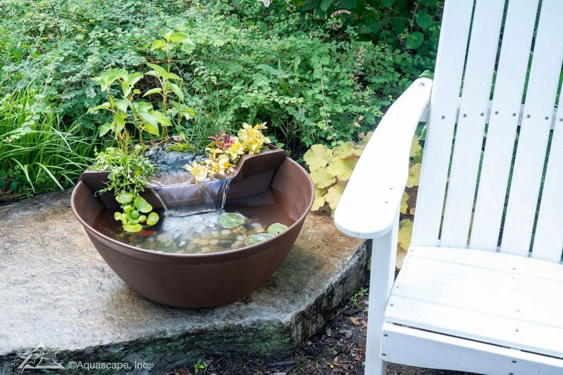 A decorative garden water feature with floating plants and flowers in a large brown container, placed on a stone surface next to a white wooden garden chair surrounded by green foliage.