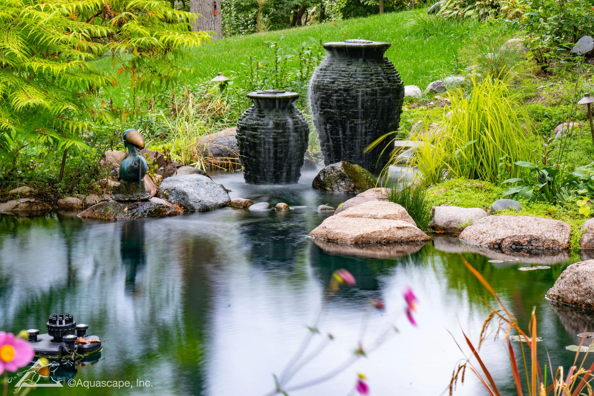 A peaceful garden pond with two large black water feature vases, a small duck sculpture, surrounded by lush green plants and rocks, with reflections in the water.