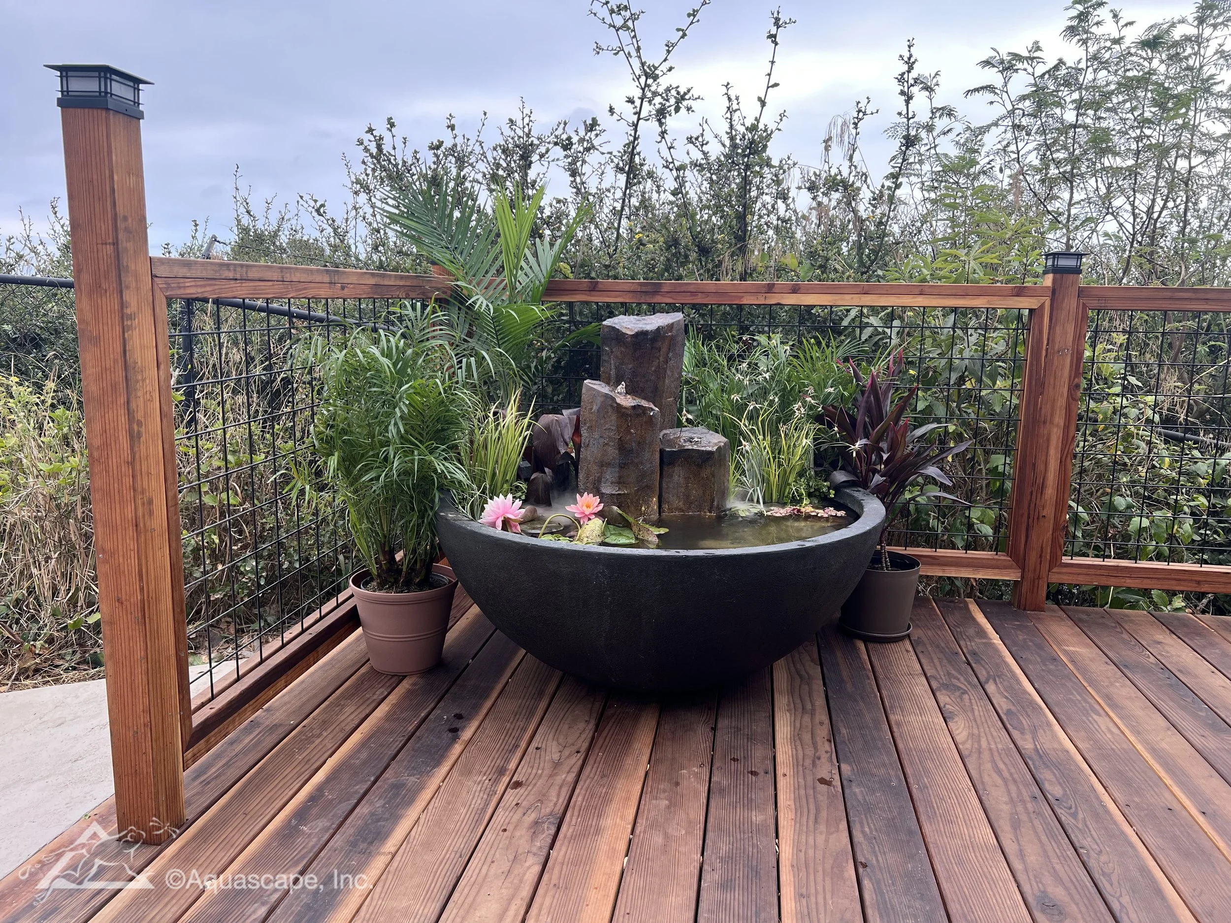 A decorative outdoor water fountain with three stone pillars surrounded by potted plants on a wooden deck with a wooden railing and greenery in the background.