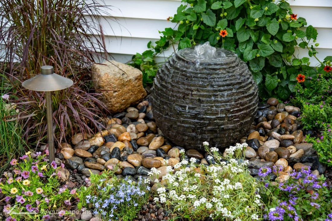 A garden water feature with a dark, spherical stone fountain releasing water, surrounded by colorful flowers, green foliage, and decorative rocks, with a small landscape light to the left.