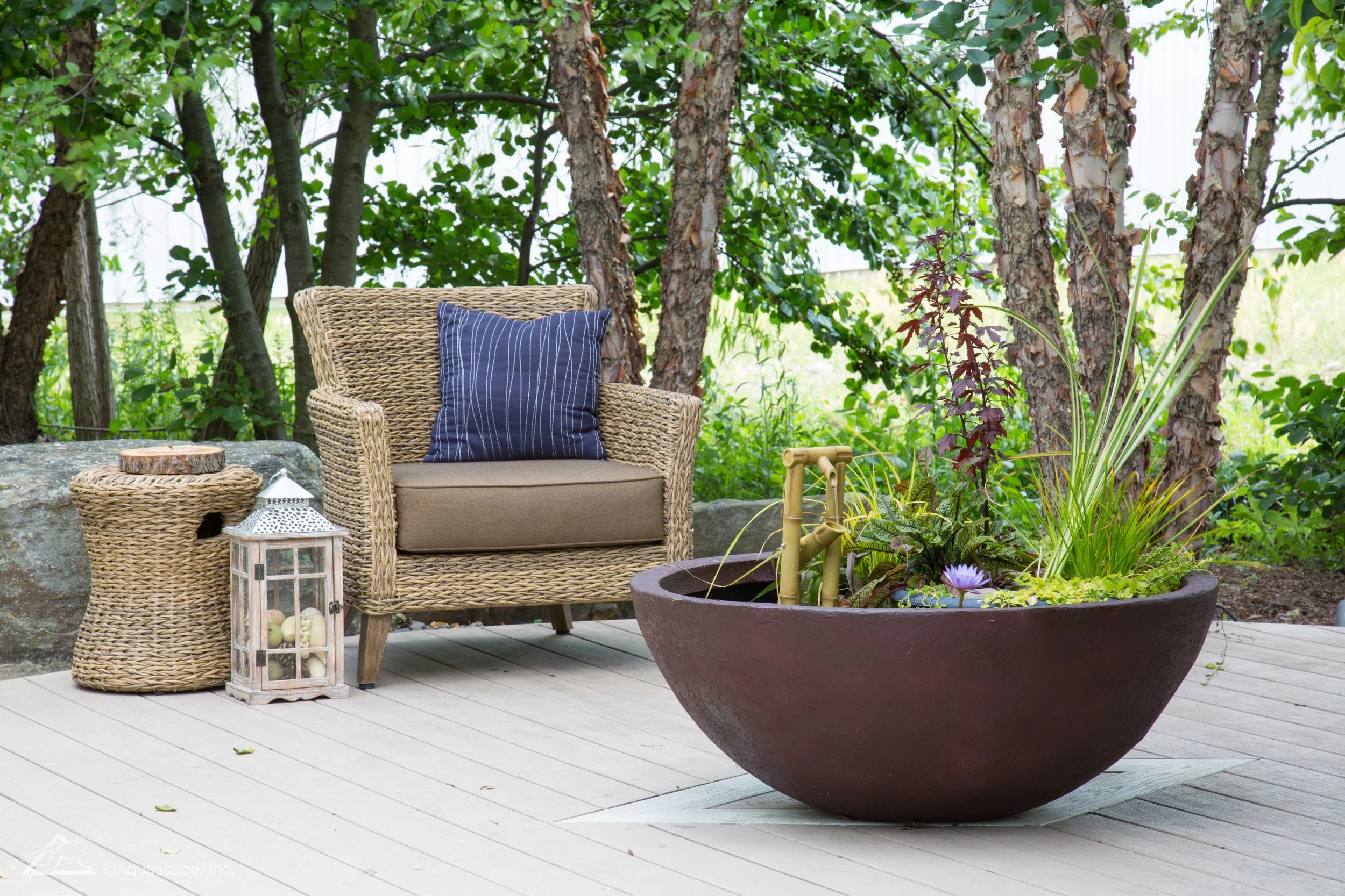A wicker armchair with a blue striped pillow on a wooden deck, next to a large planter with various plants and a small bamboo water fountain, surrounded by trees.