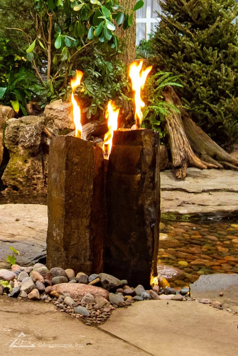 In a garden setting, there are two vertical stone pillars with flames on top, surrounded by rocks, greenery, and trees.