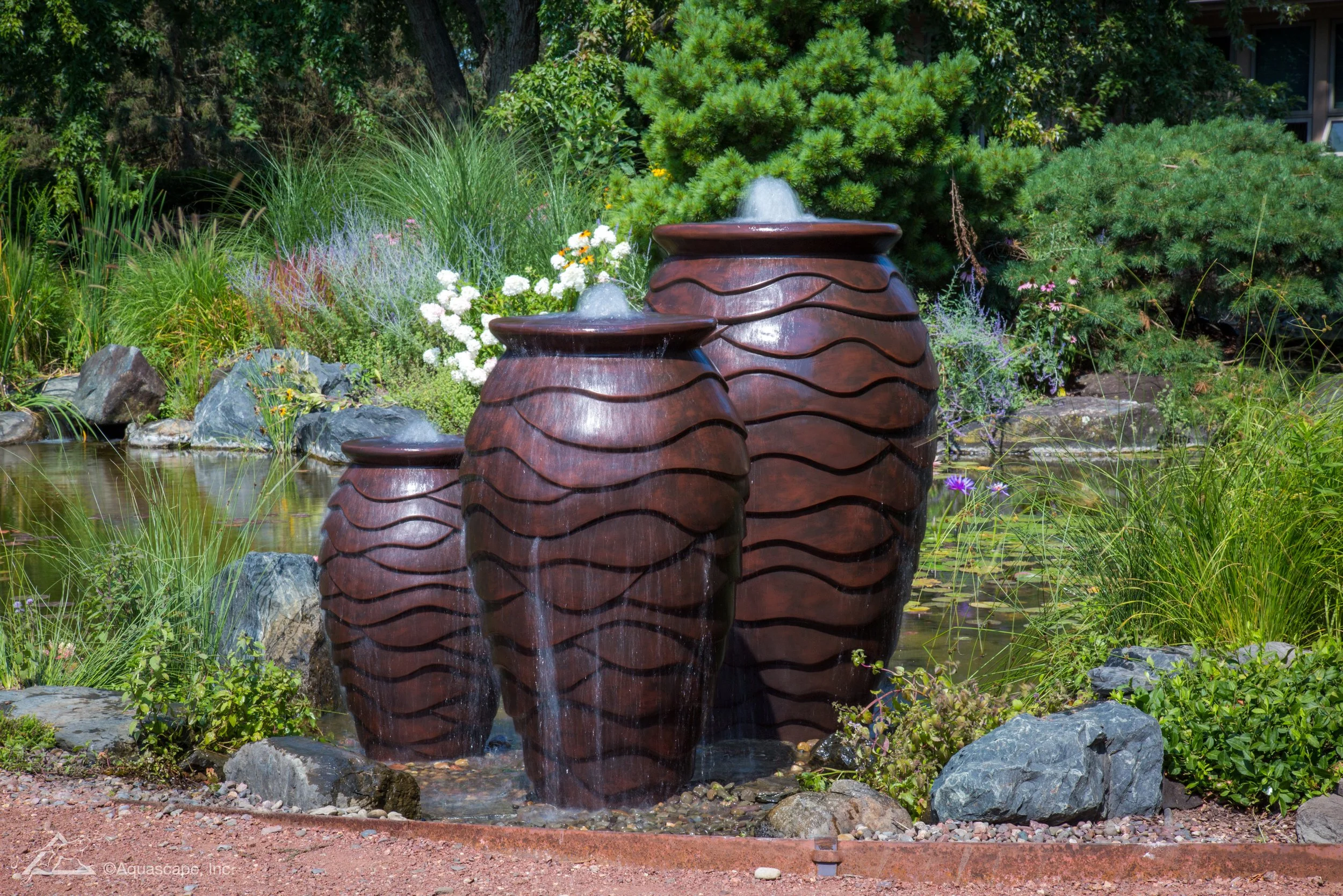 Three large decorative water fountains in the shape of vases with flowing water, placed in a garden pond surrounded by green plants, rocks, and colorful flowers.