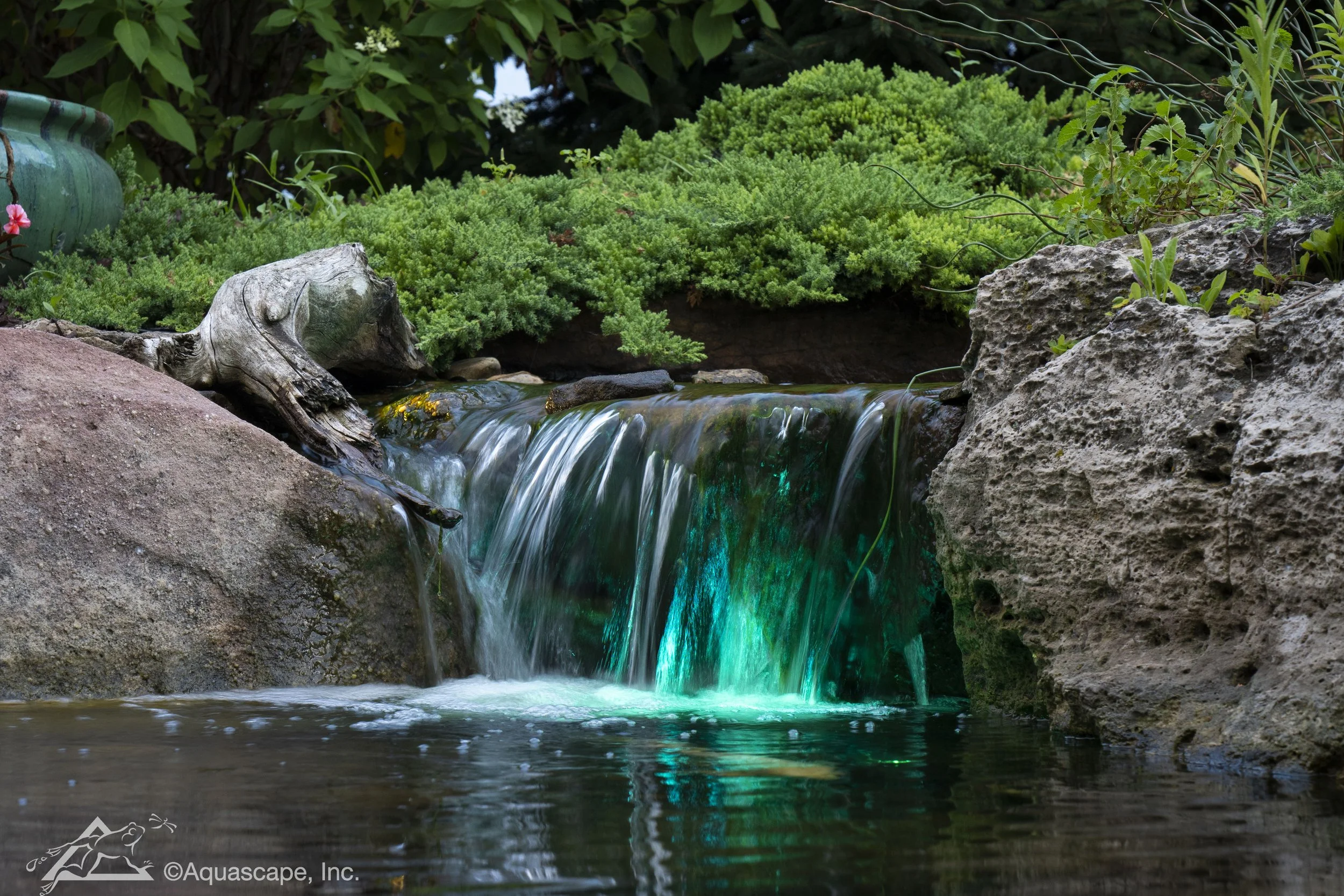 Small waterfall flowing over rocks surrounded by green bushes and plants in a garden setting.