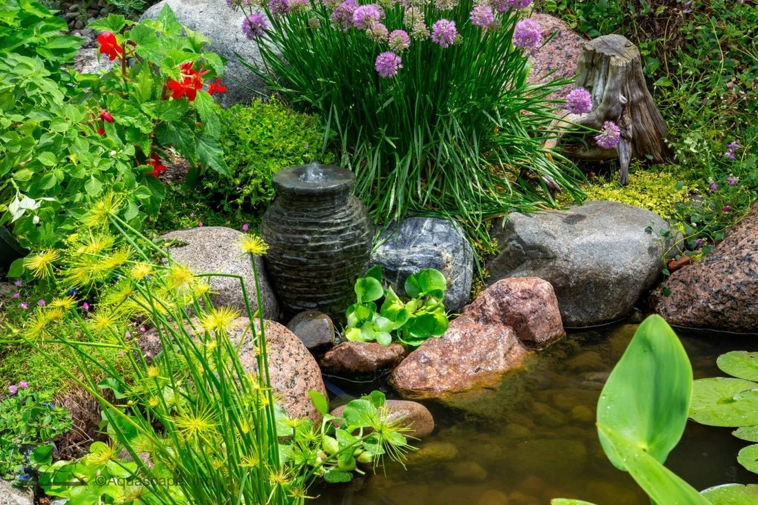 A garden pond surrounded by various green plants, colorful flowers, and rocks. There is a small water fountain, and decorative elements like a stumped tree branch with flowers.