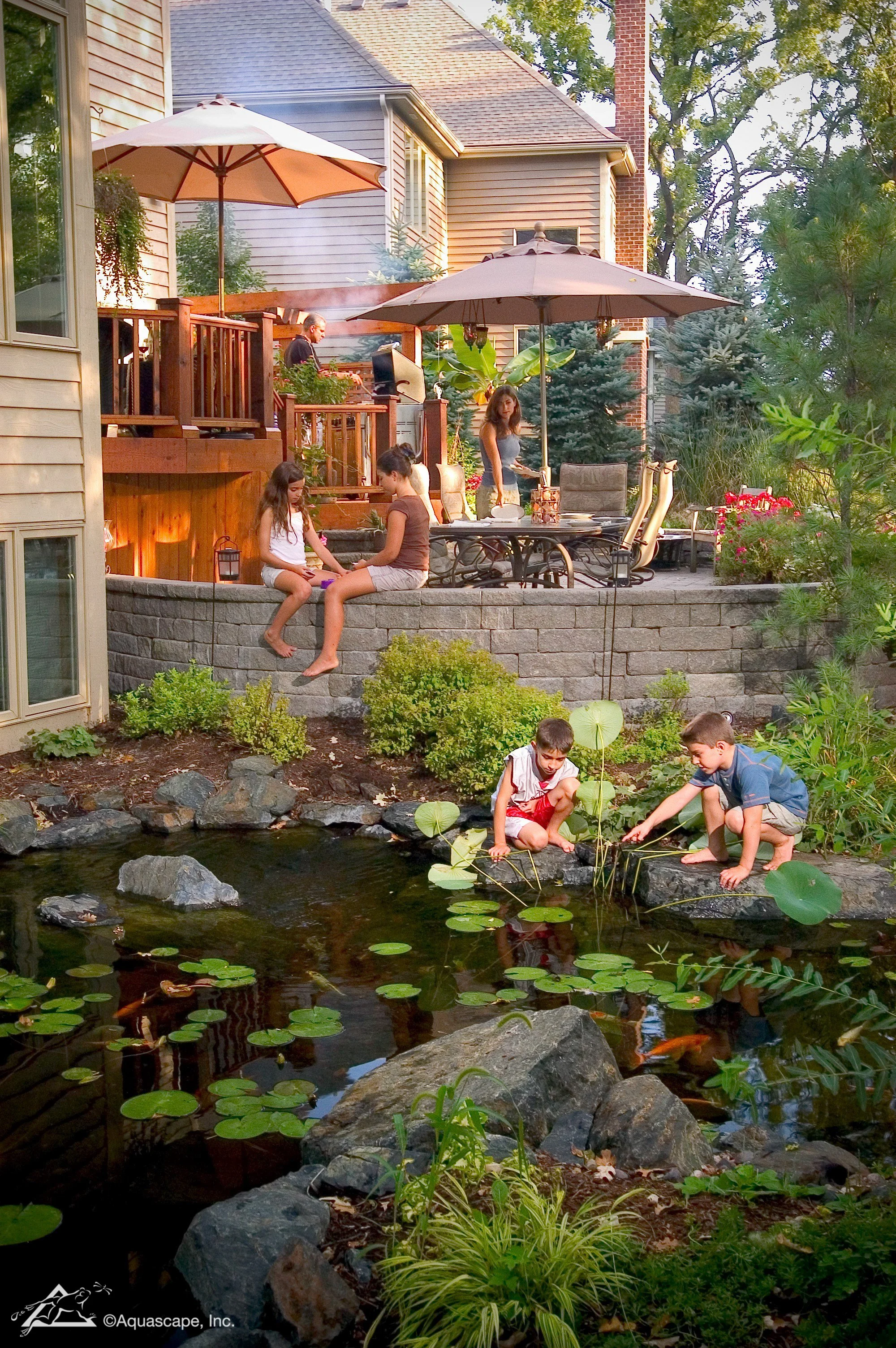 Family enjoying backyard with pond, children playing near water, outdoor dining area with umbrellas, and house in the background.