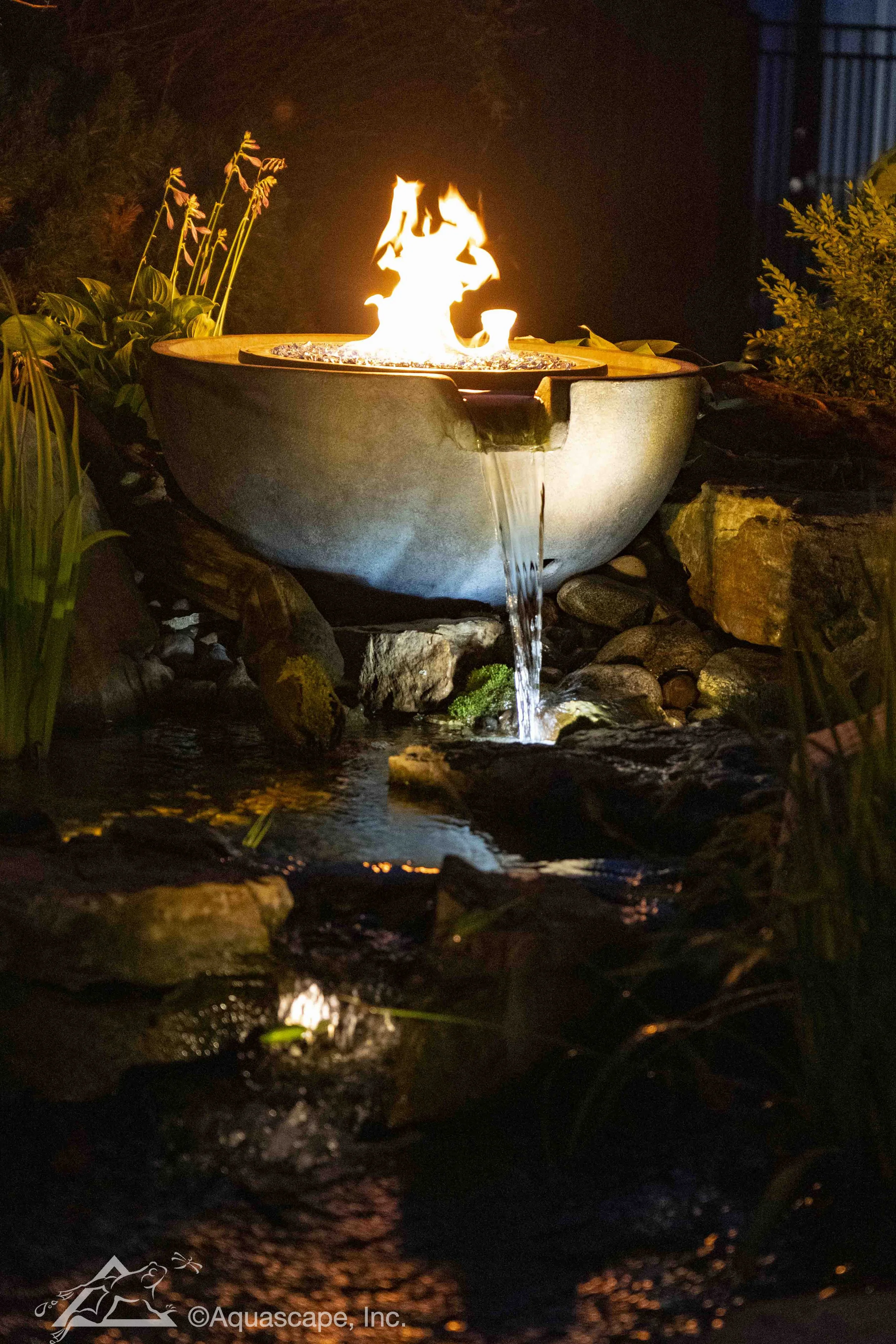 A fire feature with a simulated flame in a bowl-shaped container, water flowing over the edge into a small stream surrounded by rocks and plants at night.