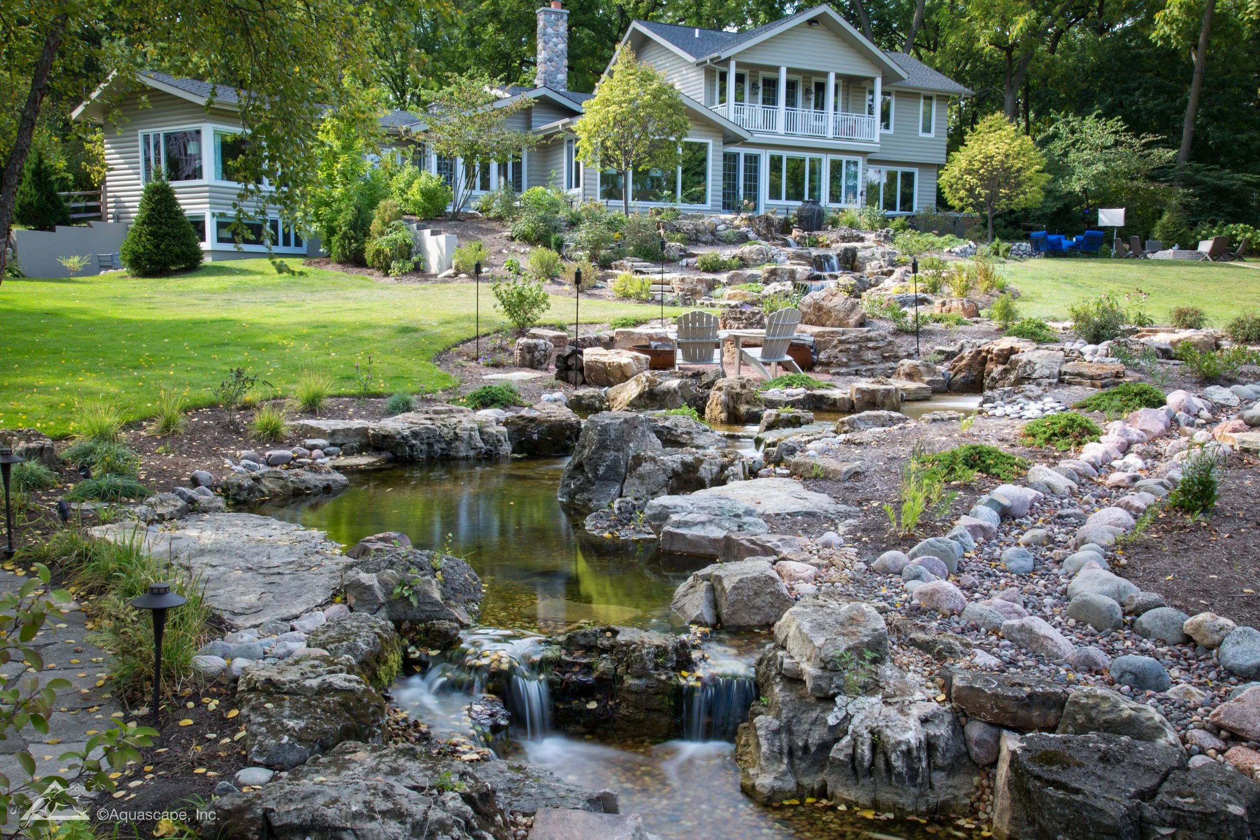 A landscaped backyard with a multi-level waterfall flowing into a pond, surrounded by rocks and plants, with Adirondack chairs nearby, and a large house in the background.