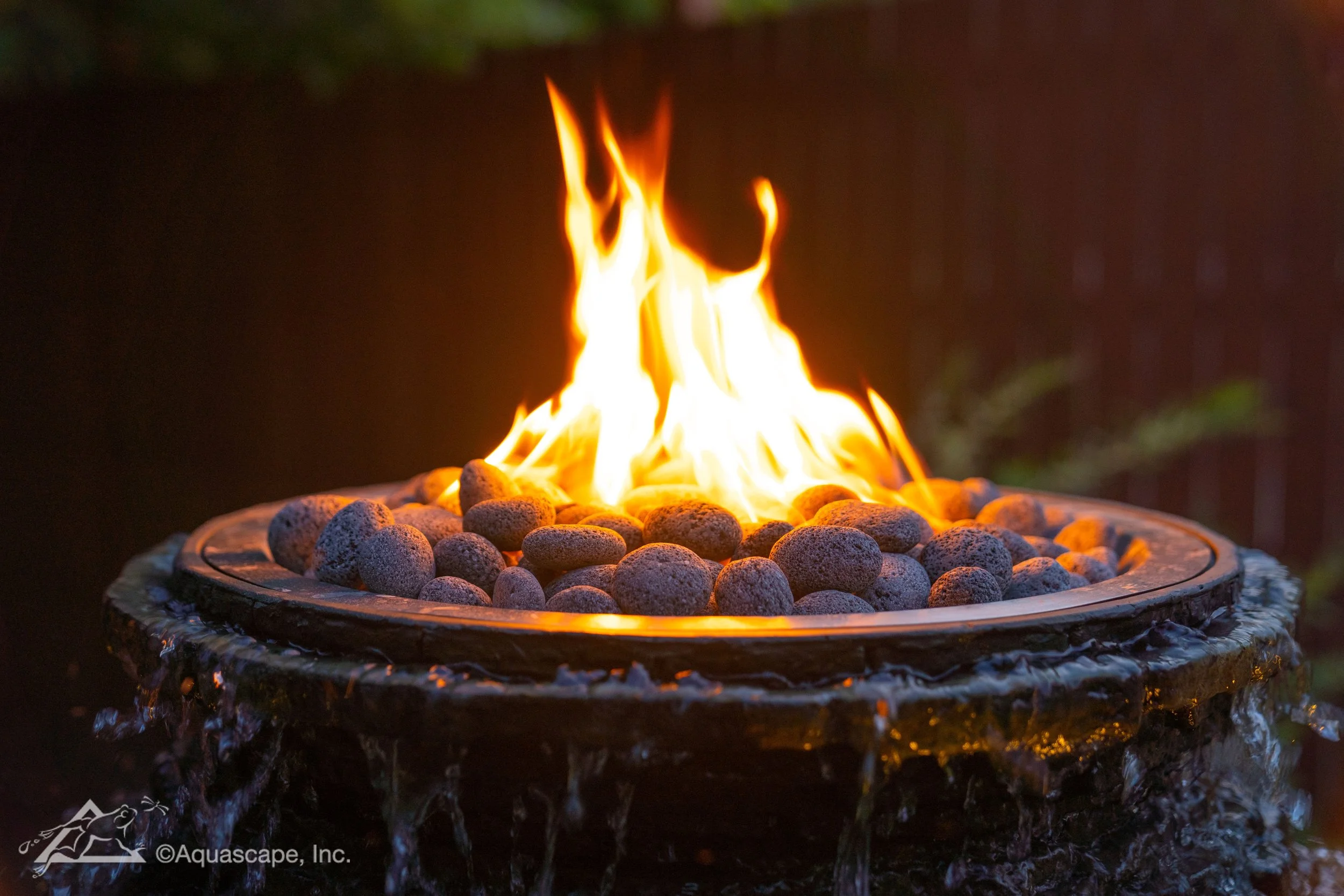 A fire burning on top of a rock and pebble setup, with flames rising from the center, surrounded by small rounded stones, outside during nighttime.