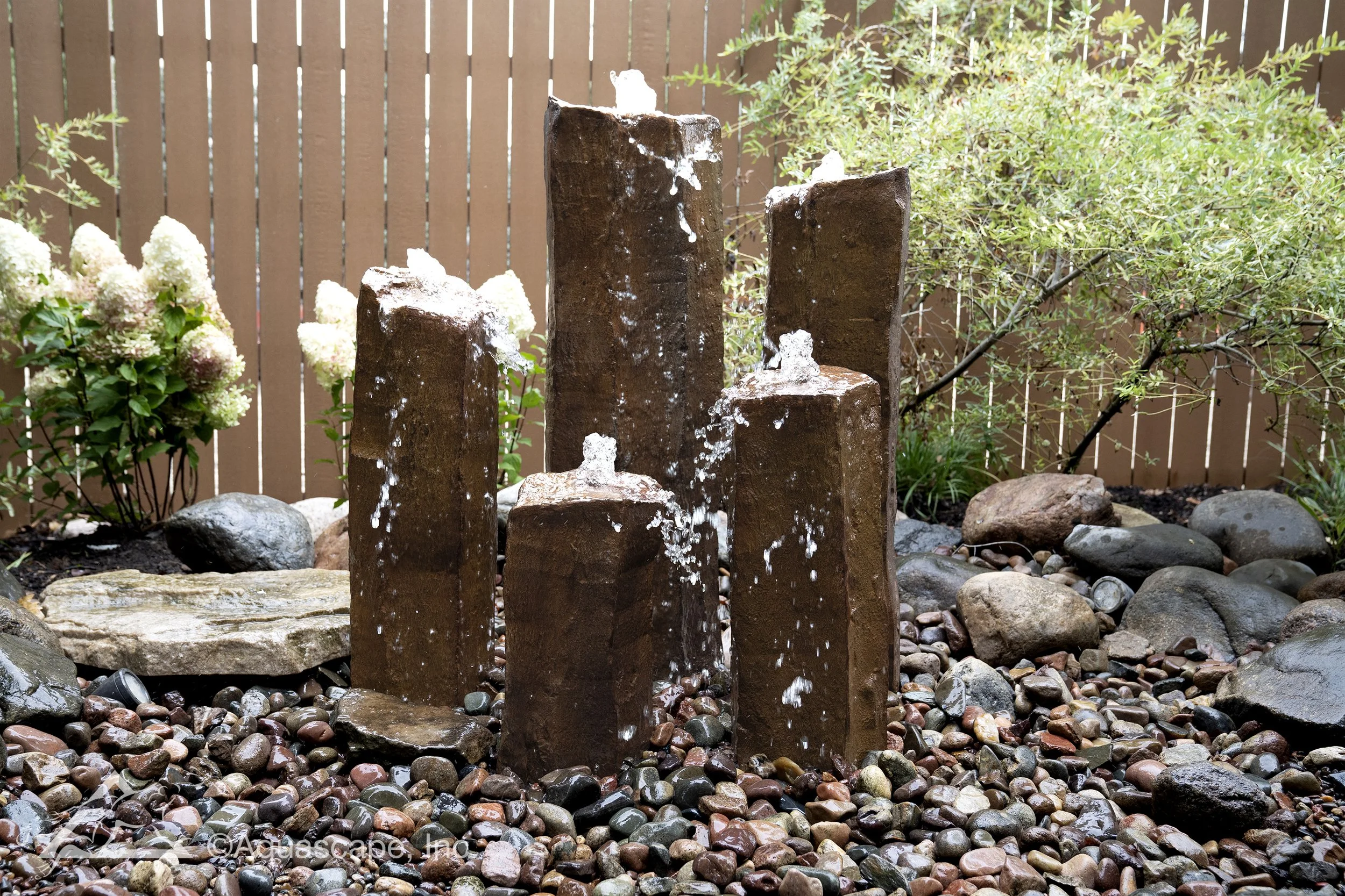 Rusty brown metal fence with a garden water fountain featuring six rectangular pillars of varying heights and water spouting from the top of each pillar, surrounded by multicolored pebbles and a bush with white flowers.