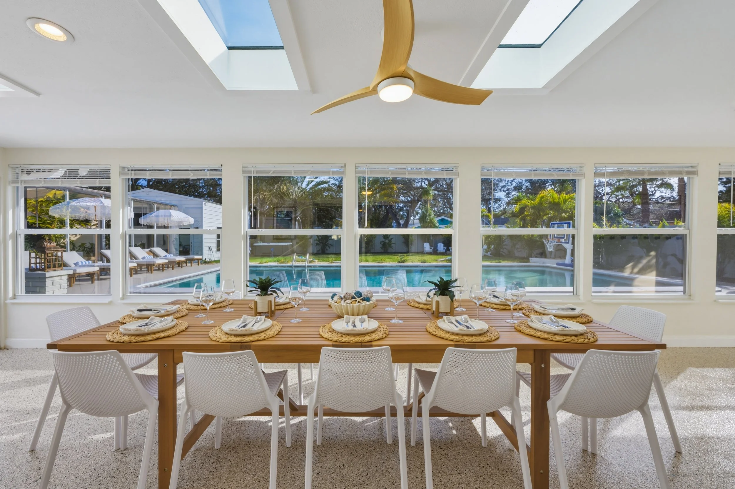 Dining table set for eight with plates, glasses, napkins, and small potted plants, overlooking a pool and backyard with trees through large windows and skylights in a bright, modern room.