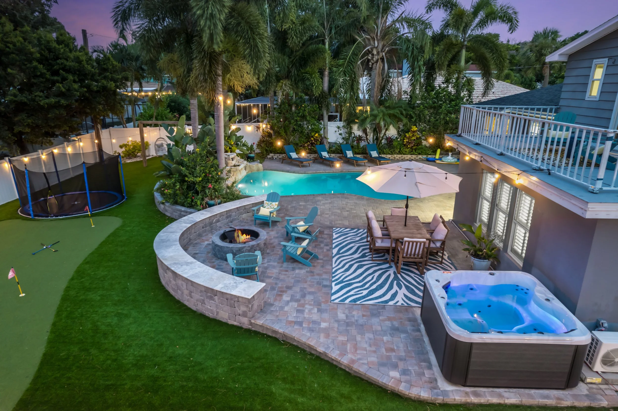 A backyard patio area at twilight with a hot tub, swimming pool, fire pit, outdoor dining, several lounge chairs, trampoline, shaded with string lights, lush trees, and a building with a balcony.