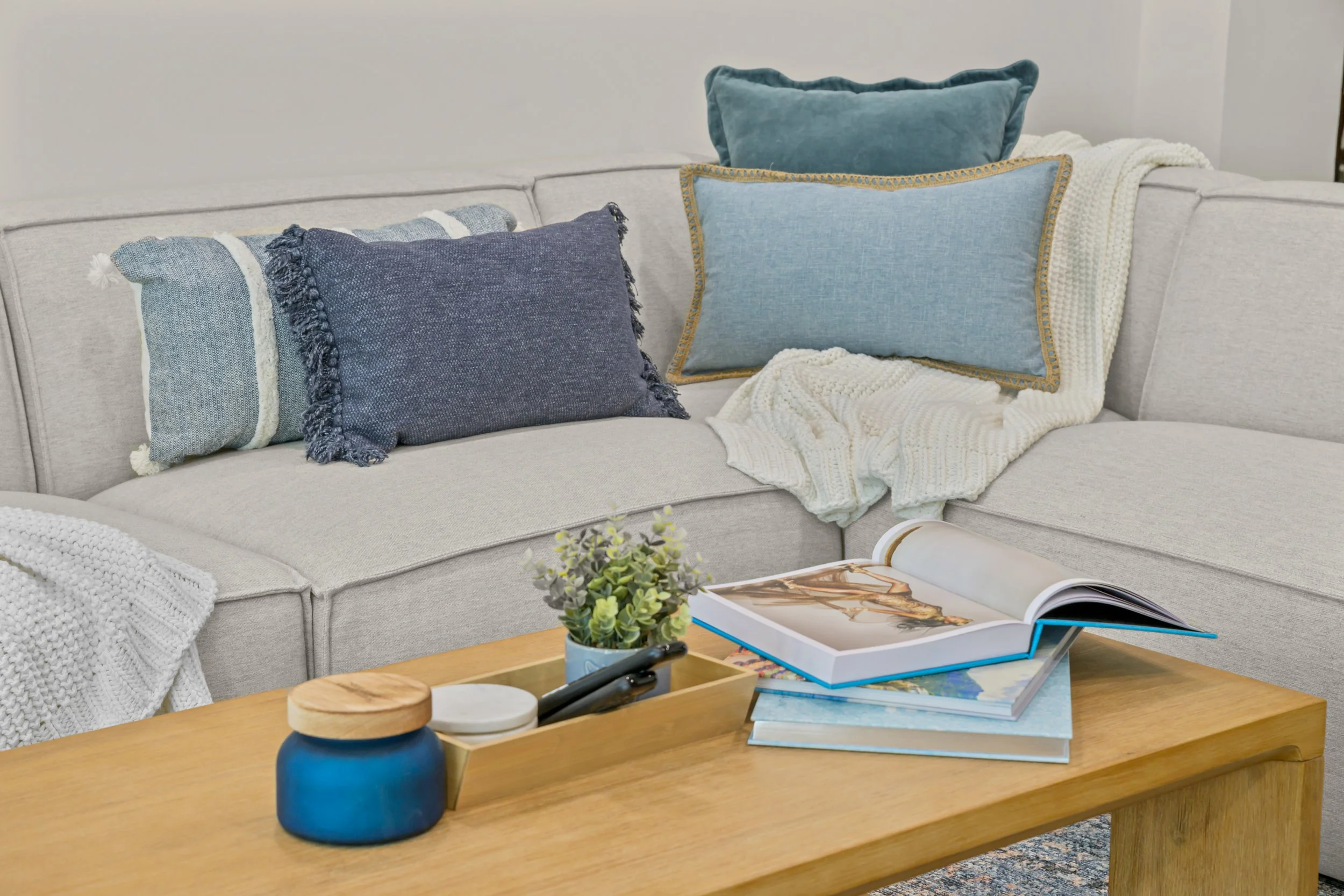 A beige sectional sofa with various blue and teal pillows, draped with a white knit blanket, in a living room with a wooden coffee table holding a small potted plant, magazines, a blue ceramic container, and a pair of glasses.