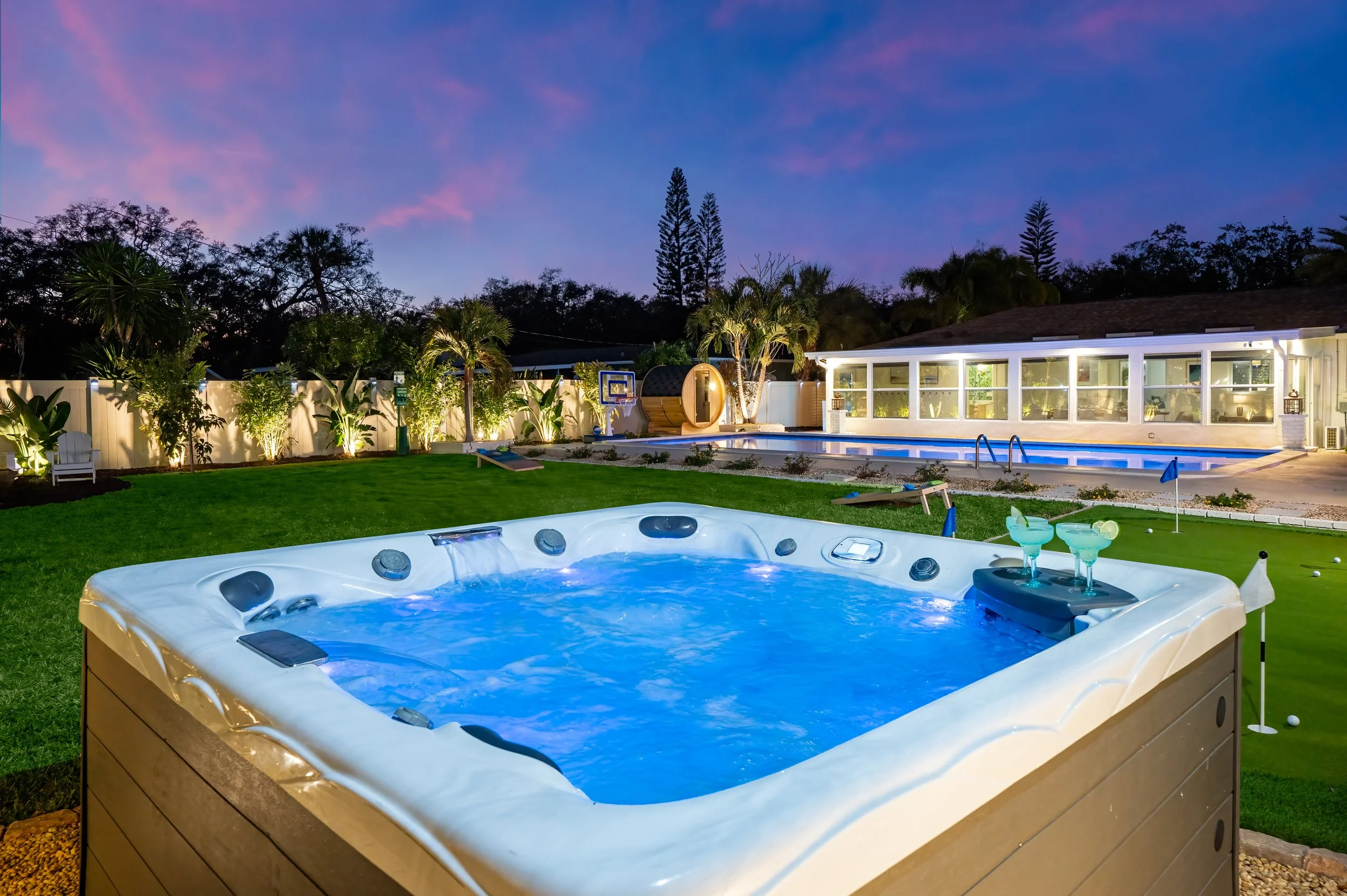 Backyard scene during dusk with spa hot tub in foreground, outdoor pool, and a lounge area, surrounded by greenery and illuminated by outdoor lights.