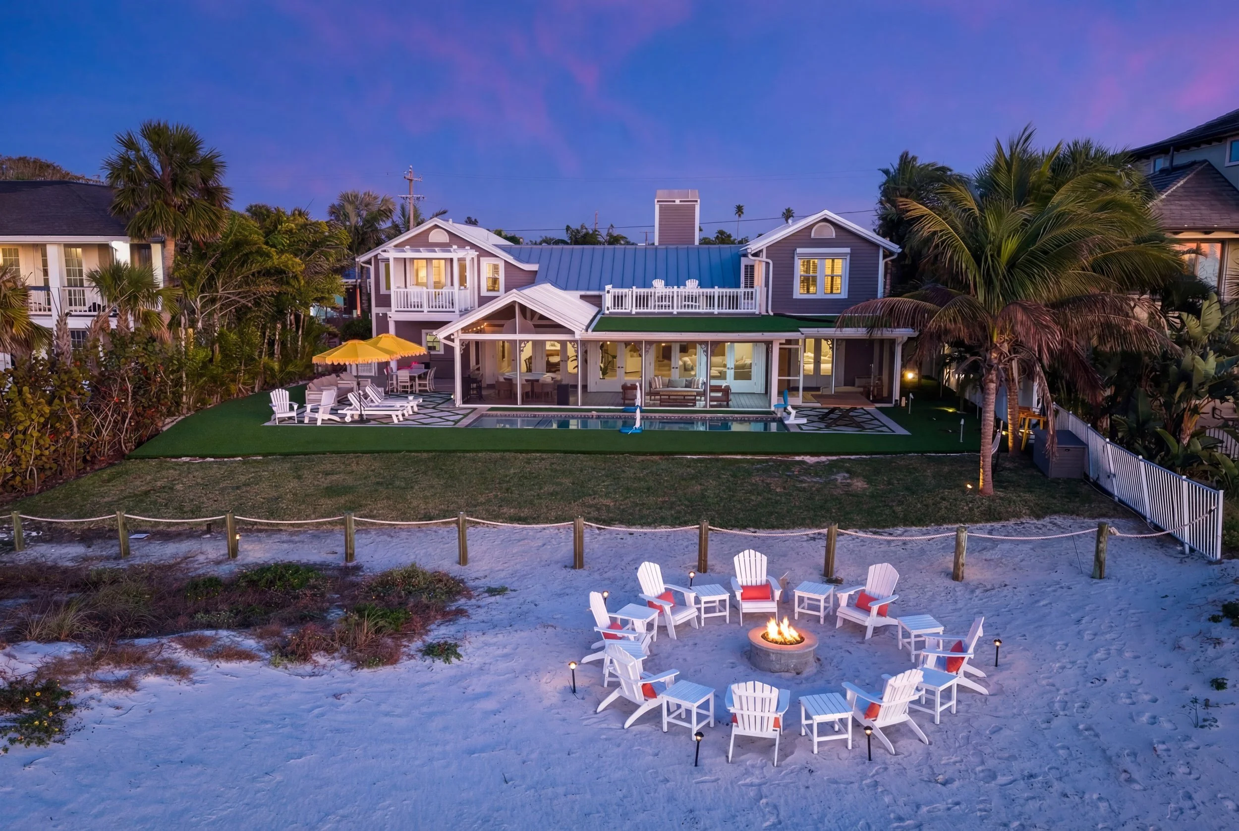 Beach house with a backyard pool, outdoor seating area, and fire pit on sandy beach under a dusk sky.