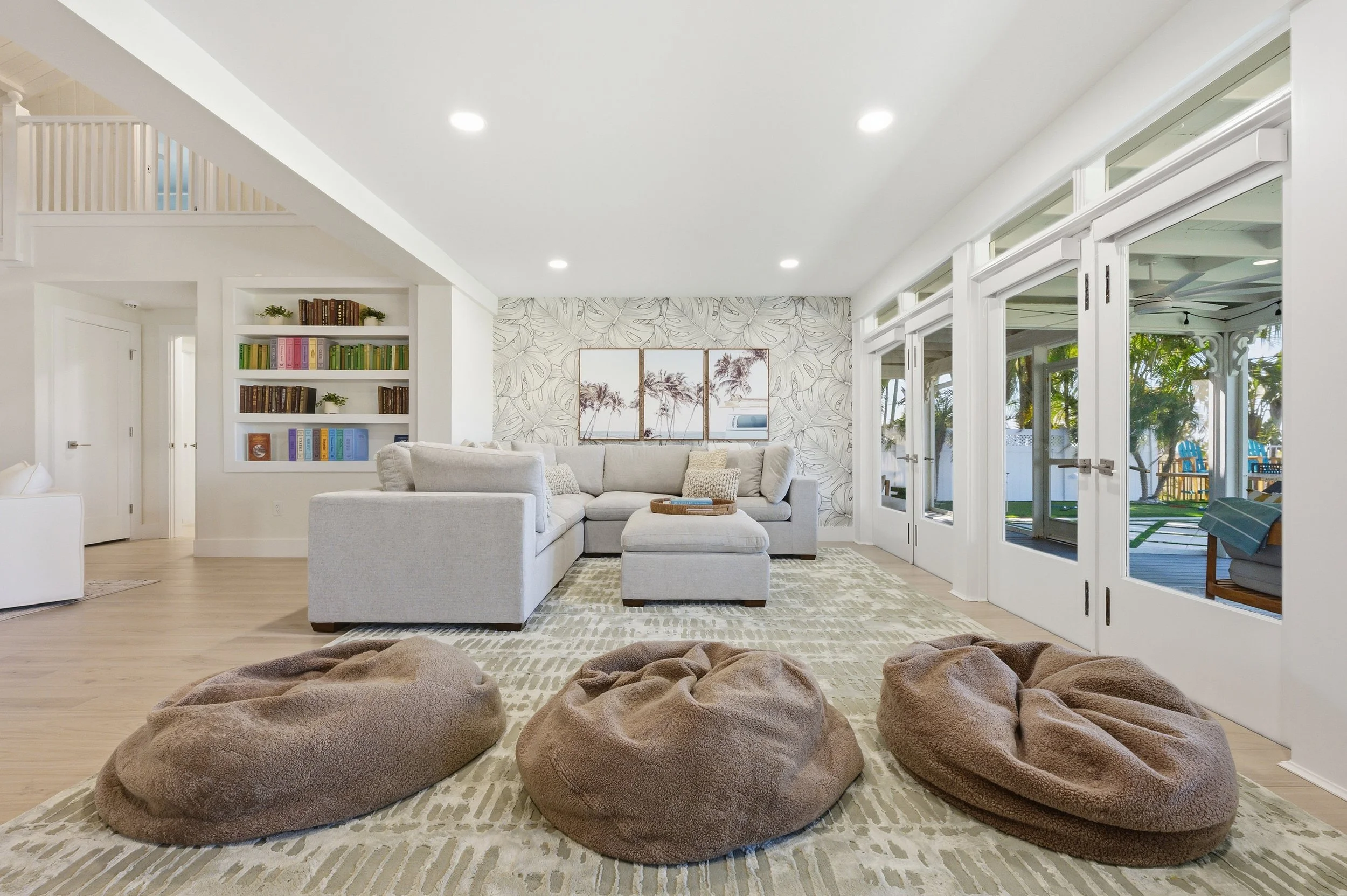 Modern living room with white sectional sofa, three brown bean bag chairs on a beige patterned rug, built-in bookshelf, and large windows with a view of outdoor patio and trees.