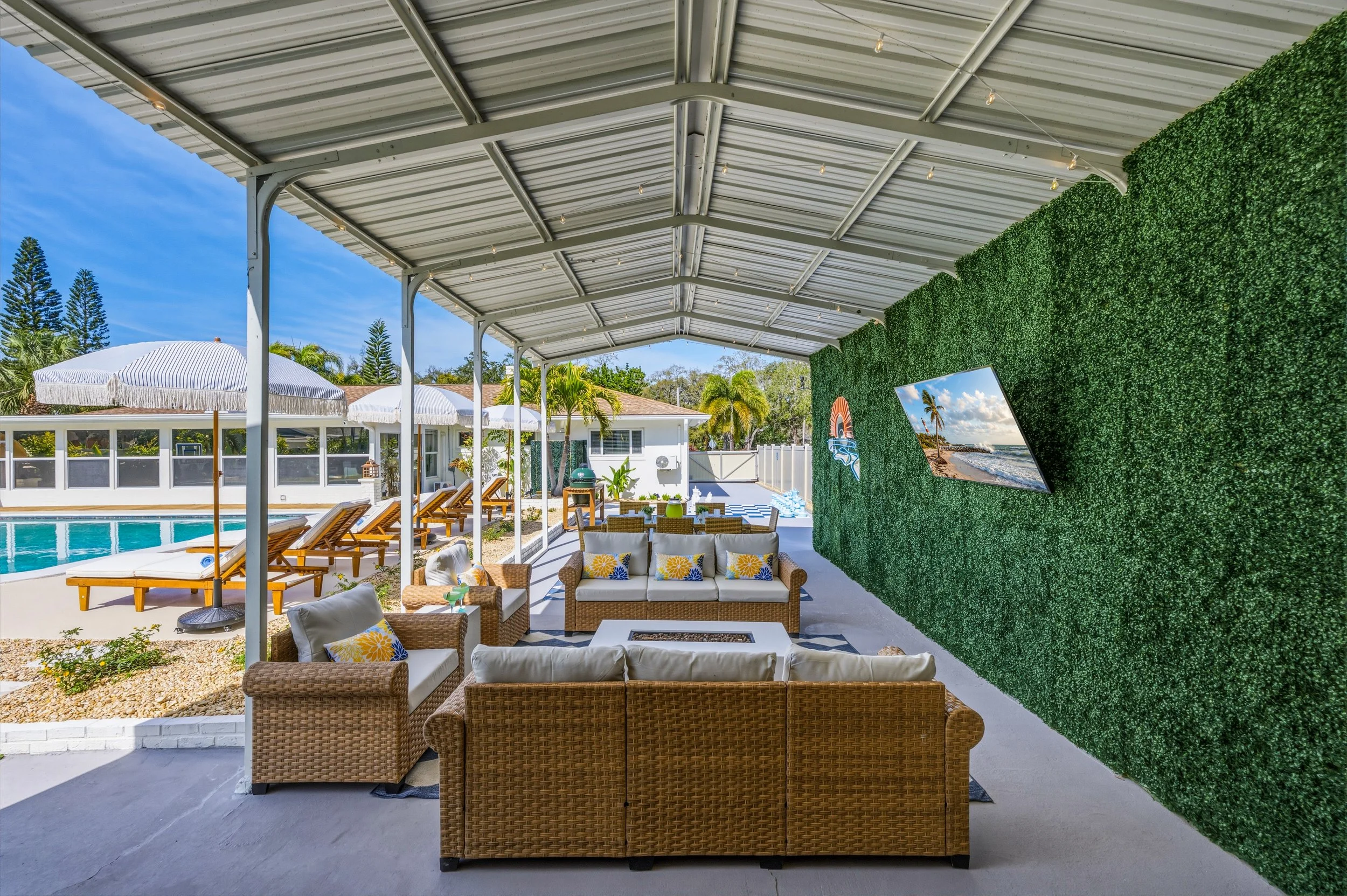 Outdoor patio area with wicker sectional sofa, chairs, and cushions facing a green artificial wall with a mounted flat-screen TV, overlooking a swimming pool with lounge chairs and umbrellas, under a metal awning in a sunny yard.